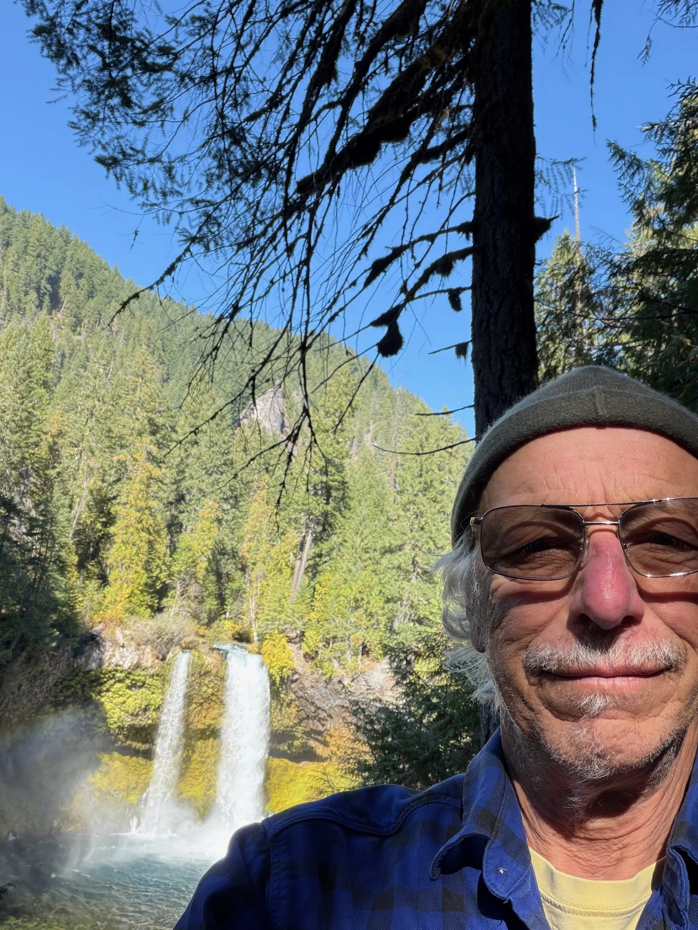 A man with gray hair, glasses, and a gray beanie taking a selfie in front of a waterfall surrounded by trees and mountainous landscape.