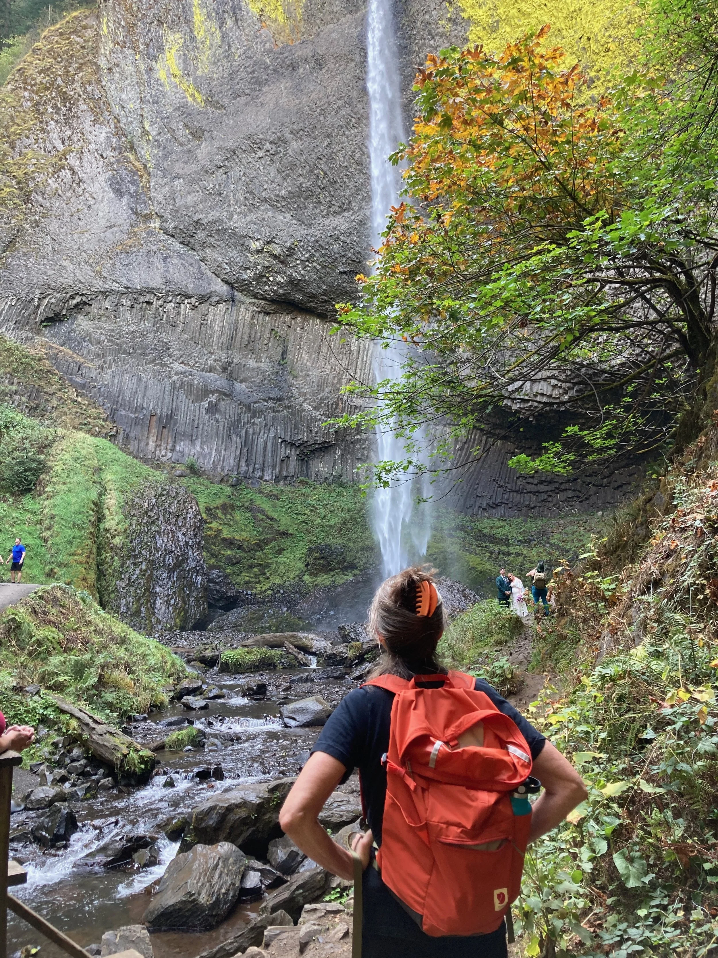 A woman with a red backpack and black shirt stands on a trail, looking at a tall waterfall cascading down a rocky cliff, surrounded by green foliage and other hikers in the background.
