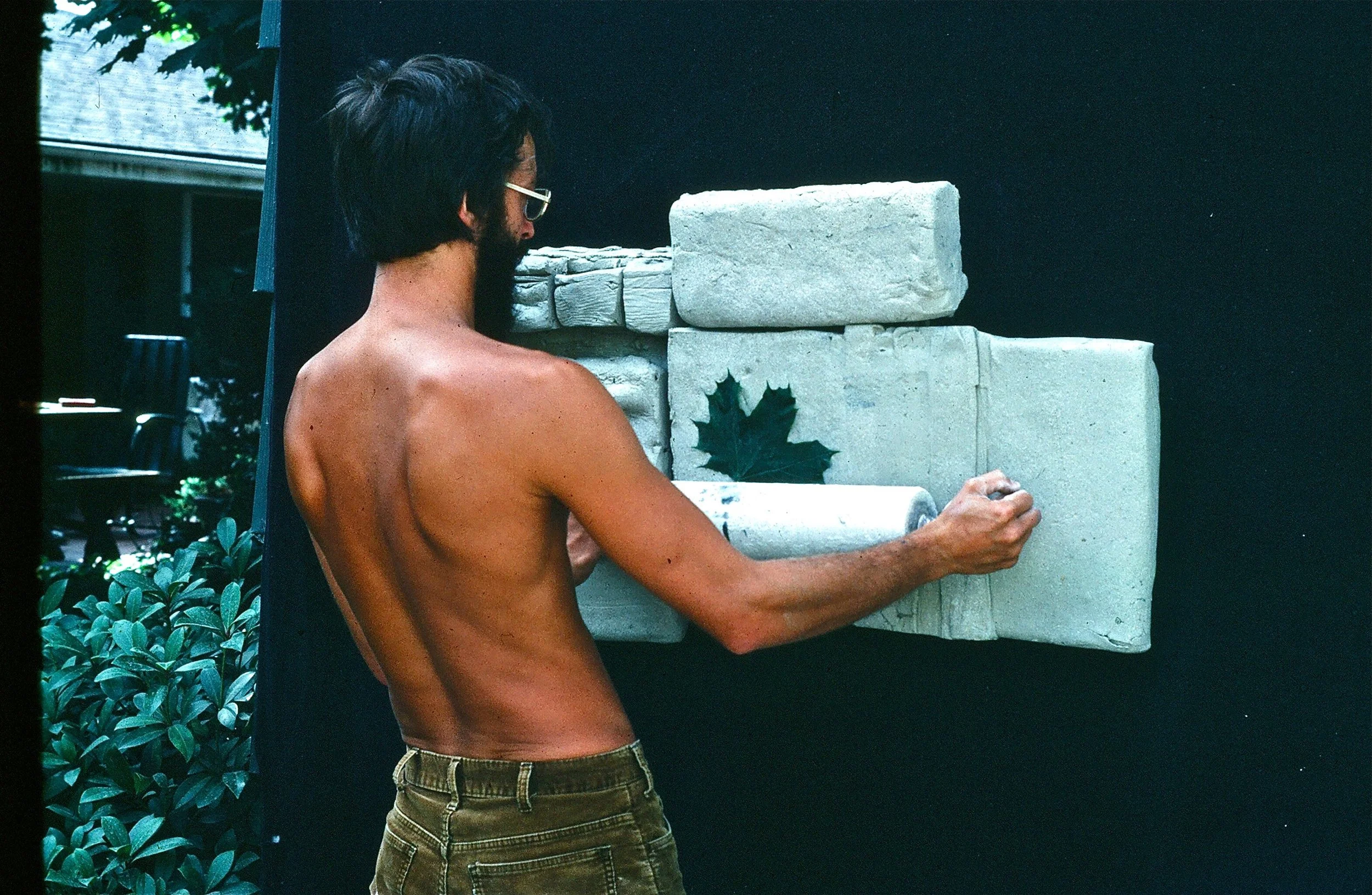 A shirtless man with glasses and dark hair is standing outdoors, facing a black backdrop, and touching a sculpture made of multiple stone blocks, some with carved details, with a green leaf painted or attached on one of the stones. There are green bu