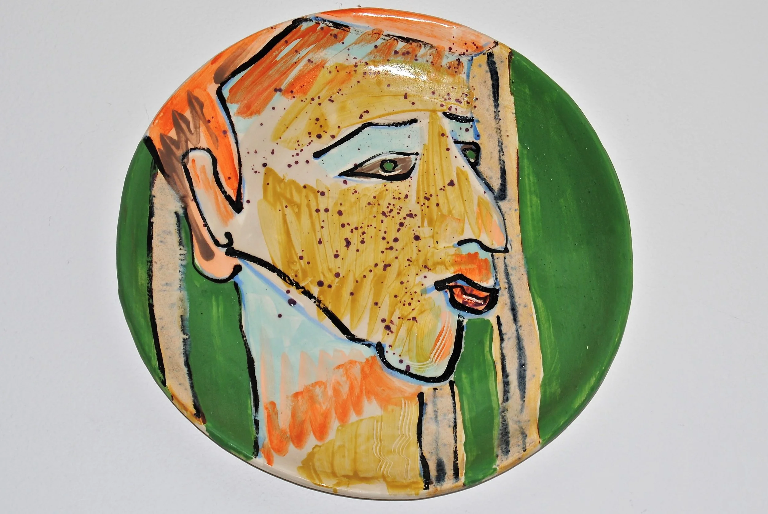 A ceramic plate with a colorful abstract portrait of a person's face, featuring yellow, tan, orange, green, blue, and black brushstrokes.