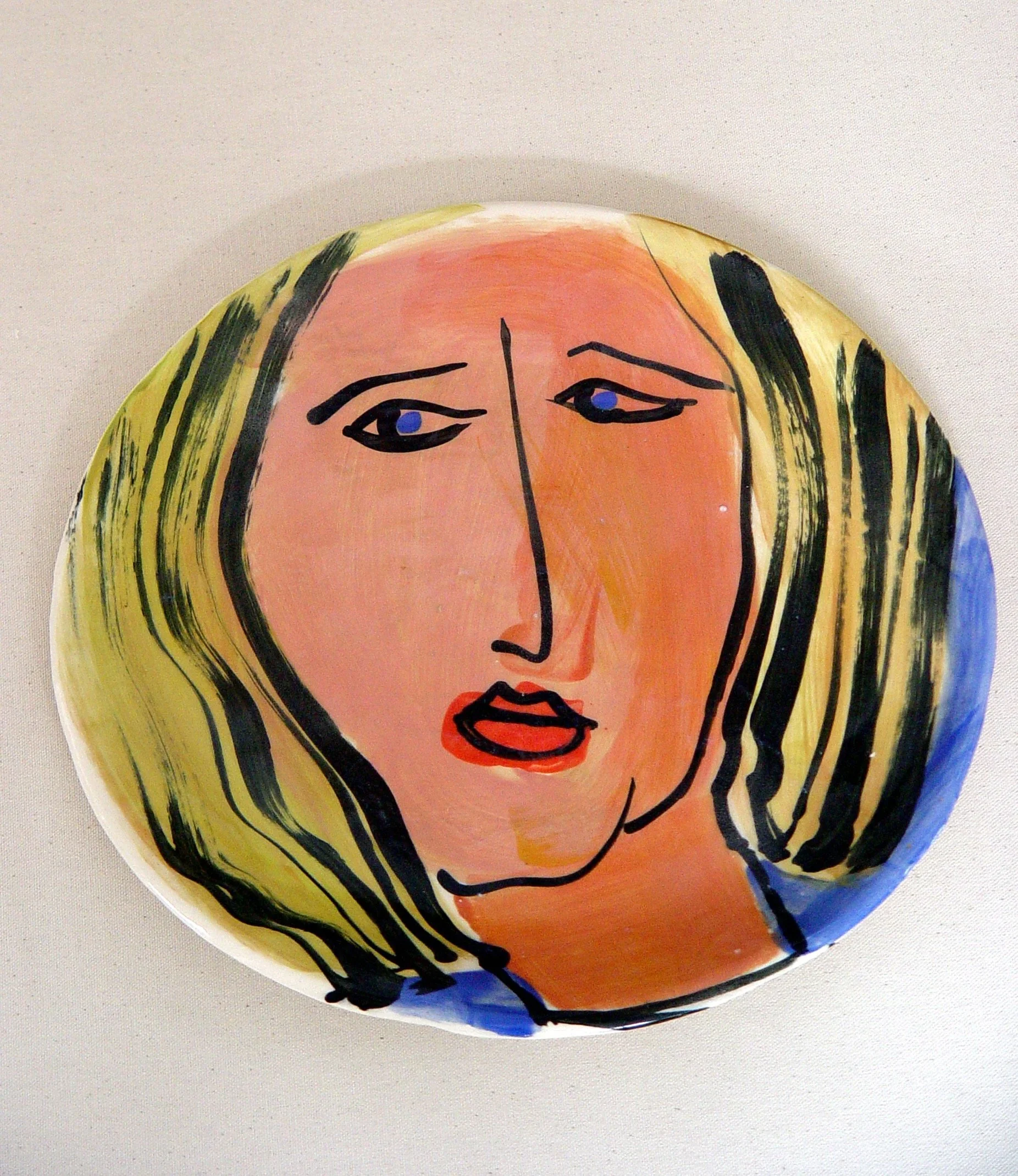 A painted ceramic plate featuring an abstract portrait of a woman with blonde hair, painted with bold black lines, blue eyes, and red lips.