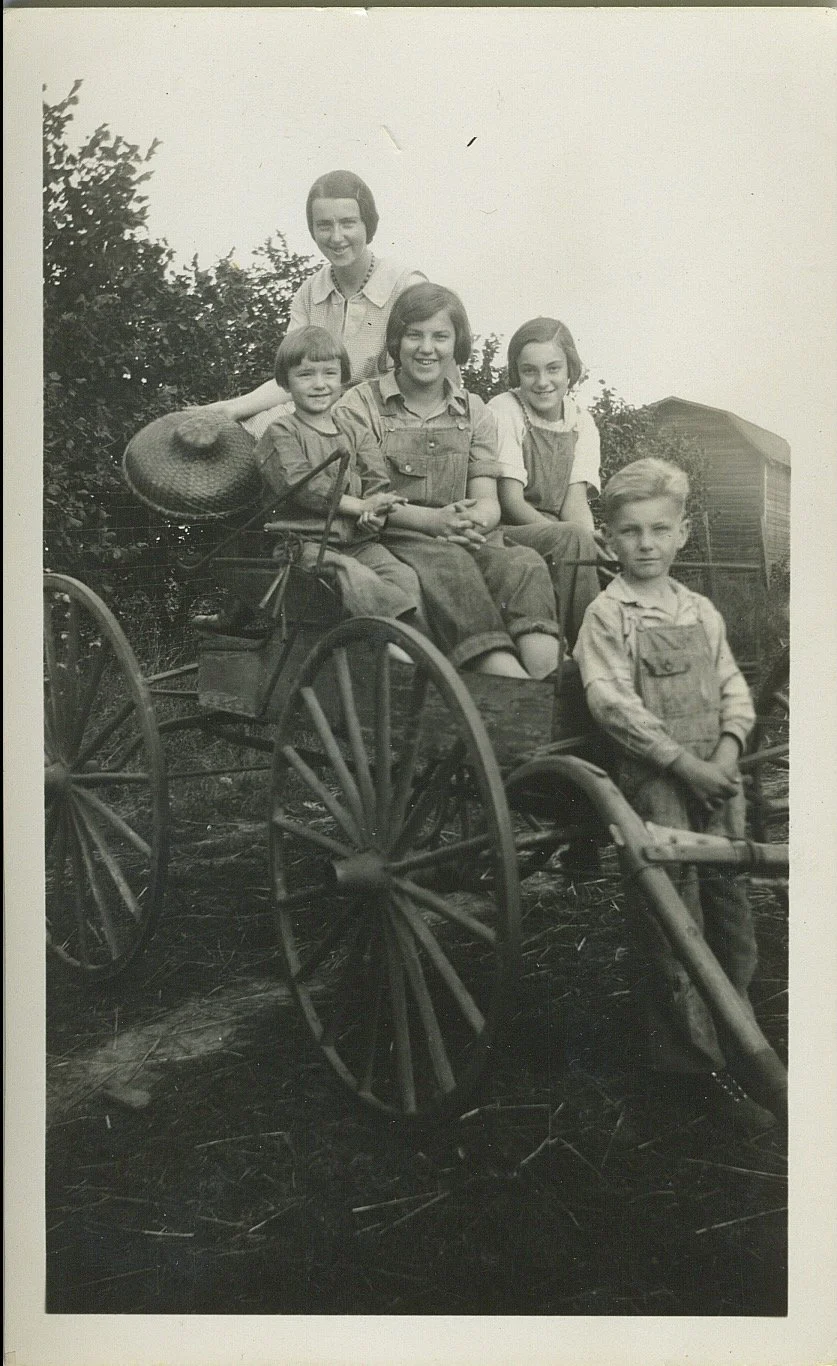 Black and white photograph of four children sitting on a wooden cart attached to a tricycle with large wheels, outdoors with trees and a building in the background.