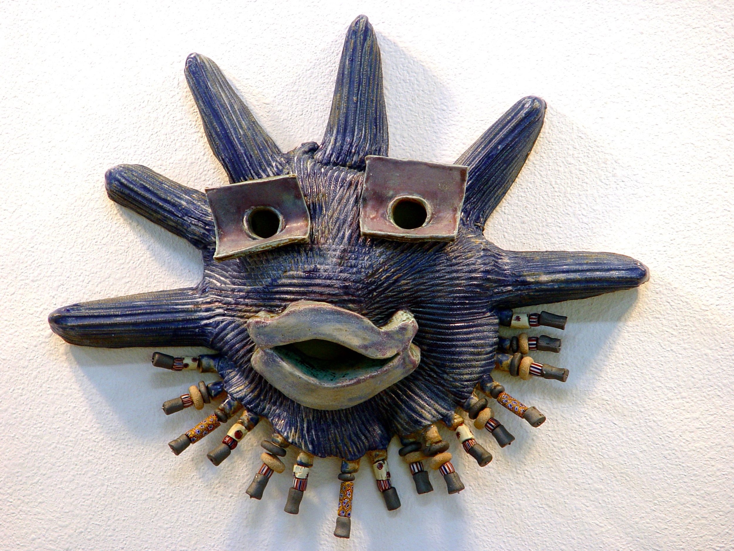 A decorative wall sculpture of a face with spiky hair, glasses, and a large open mouth, adorned with various small objects like beads and tubes.
