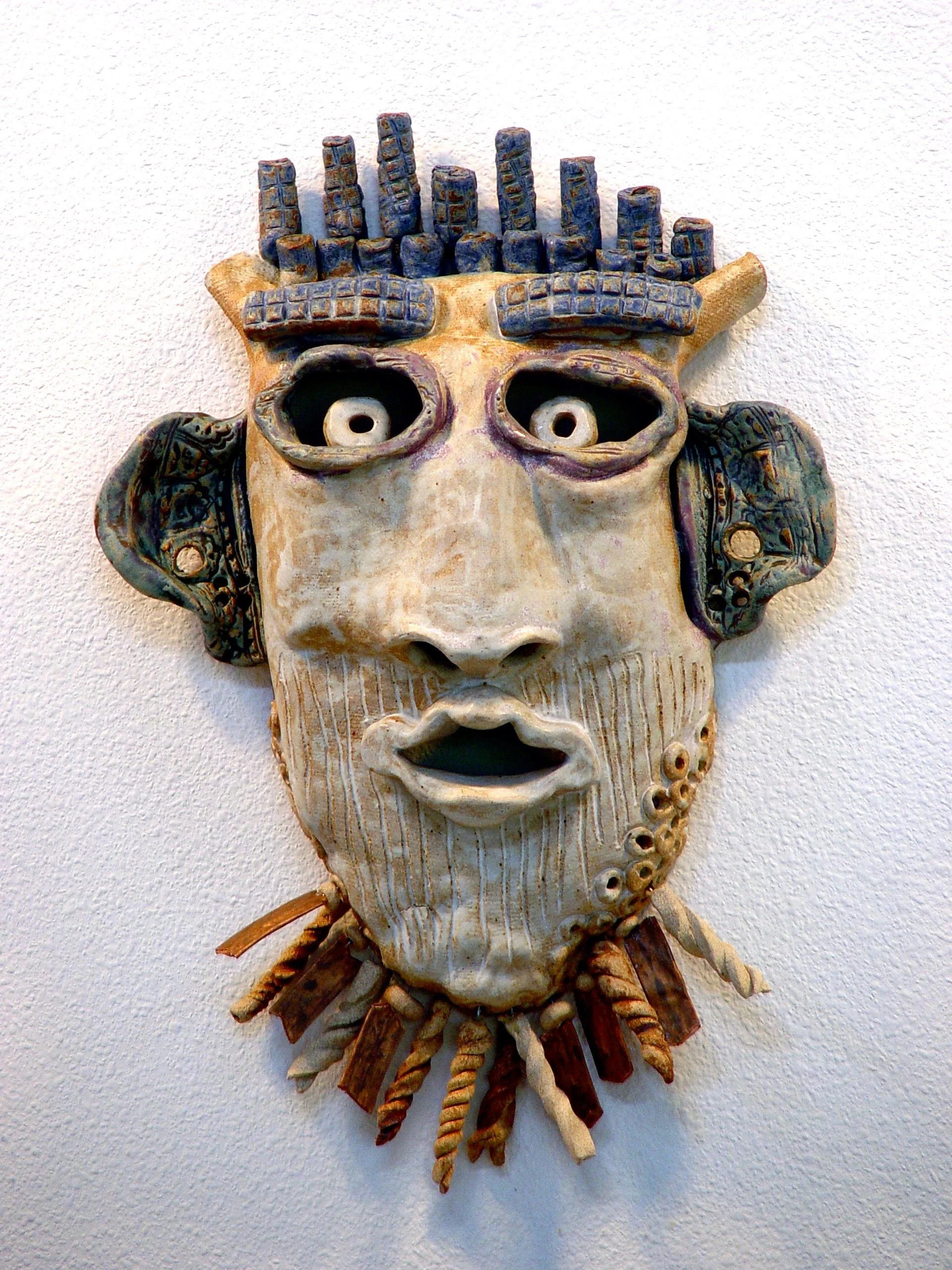 A colorful, artistic wall mask featuring a face with exaggerated features, large eyes, and textured hair made of various materials.