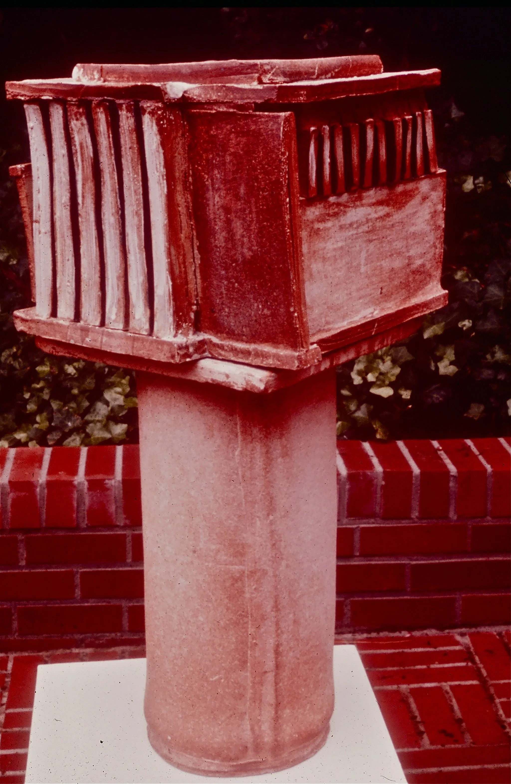 A red-painted mailbox mounted on a cylindrical pedestal, placed against a brick wall and shrubbery background.