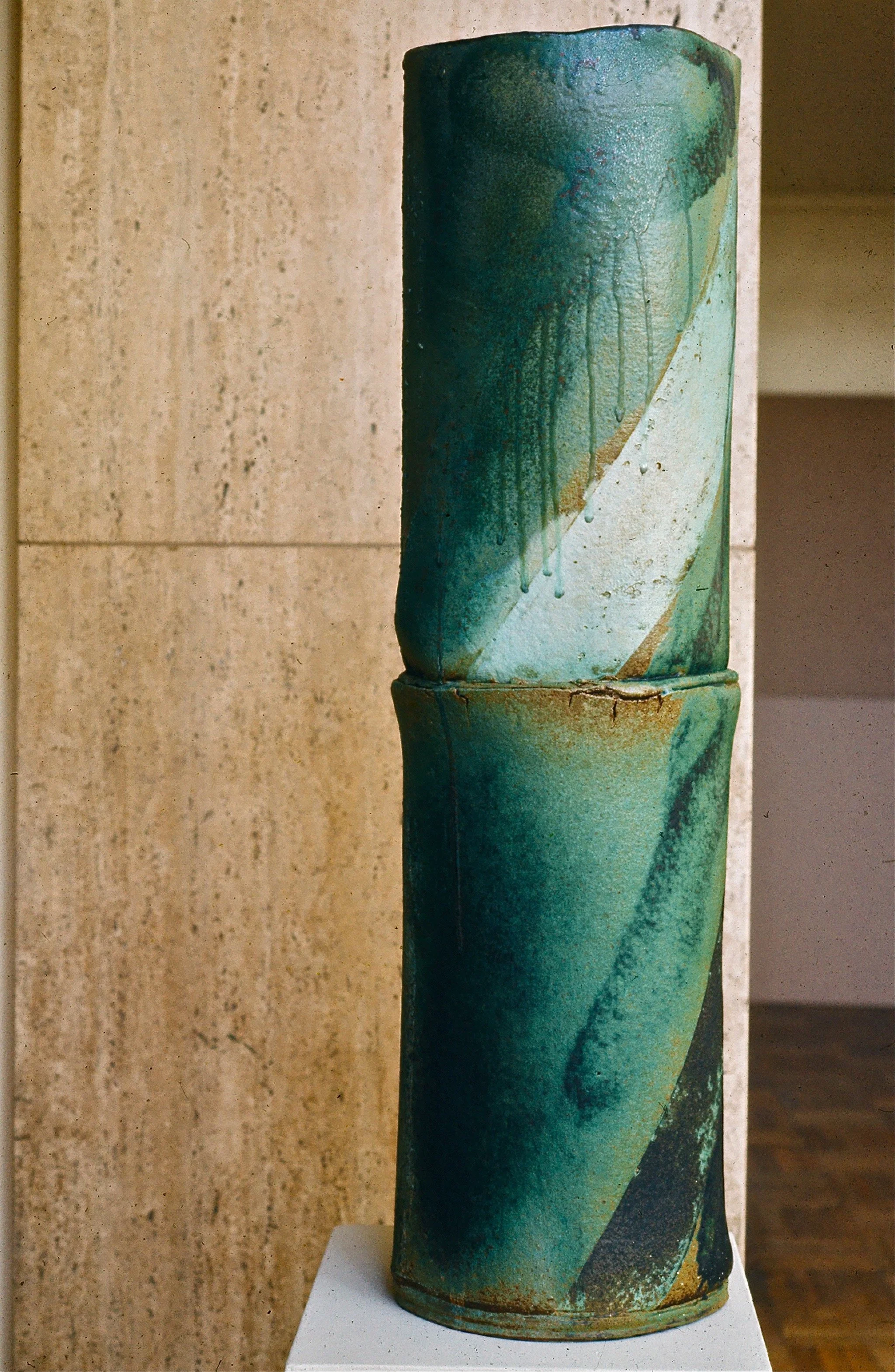 Ceramic sculpture with abstract green and blue glaze drips, stacked in two sections, on a white pedestal against a tan stone wall.