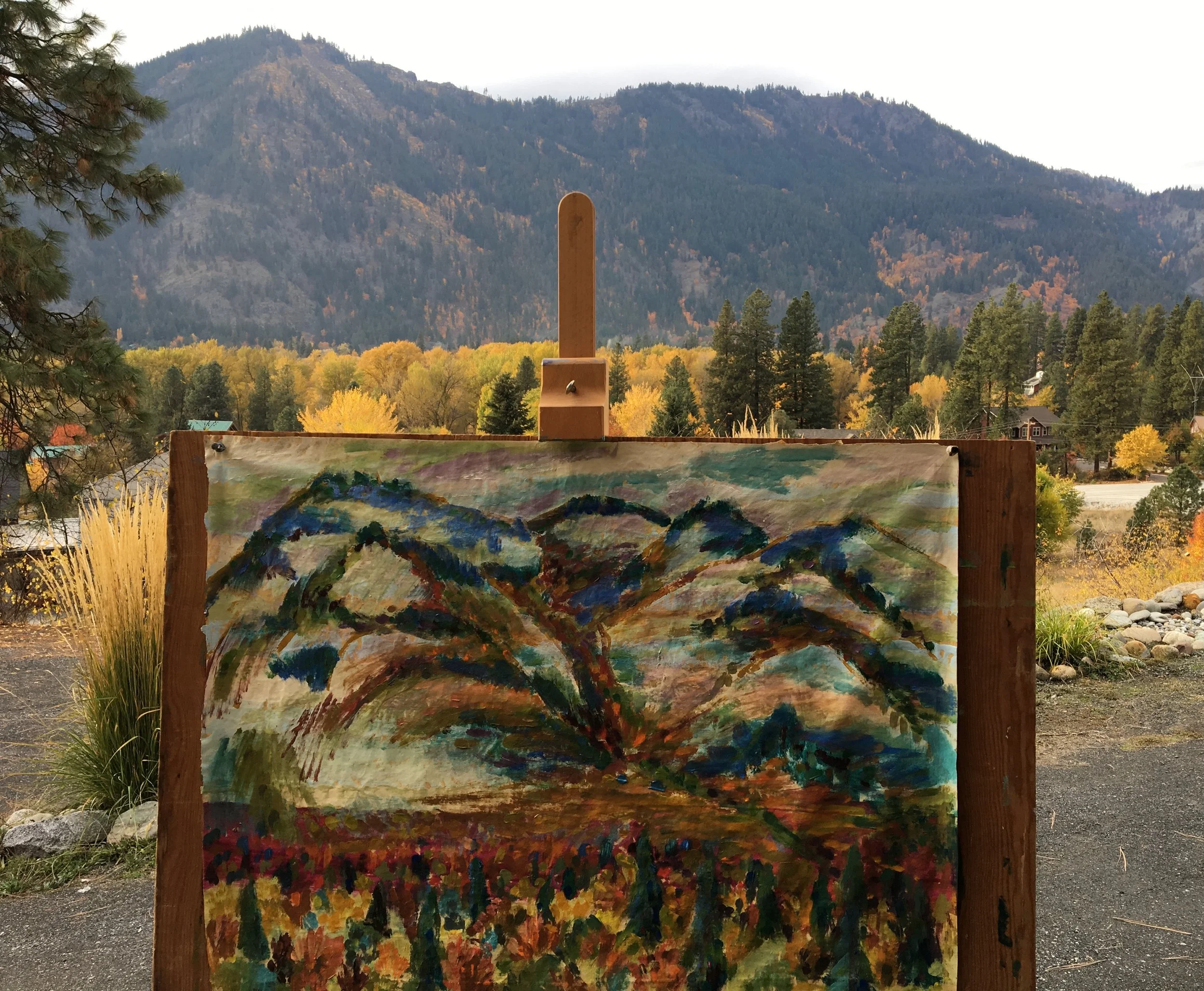 An outdoor landscape with a mountain range in the background, trees with fall foliage, and an artist's easel with a colorful, abstract painting of mountains and landscape in the foreground.
