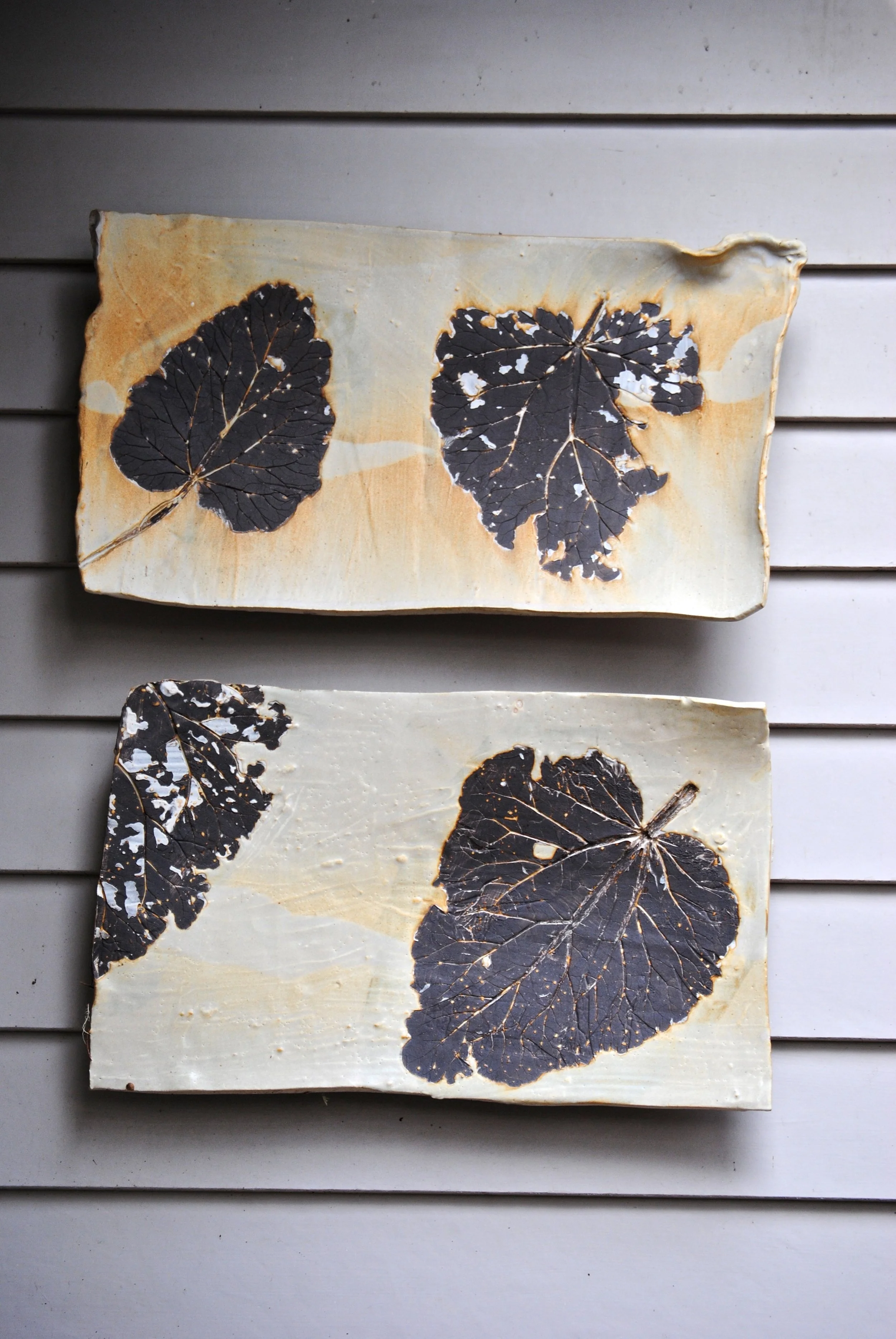 Two rectangular ceramic tiles with dark leaf patterns, placed on horizontal wooden slats.