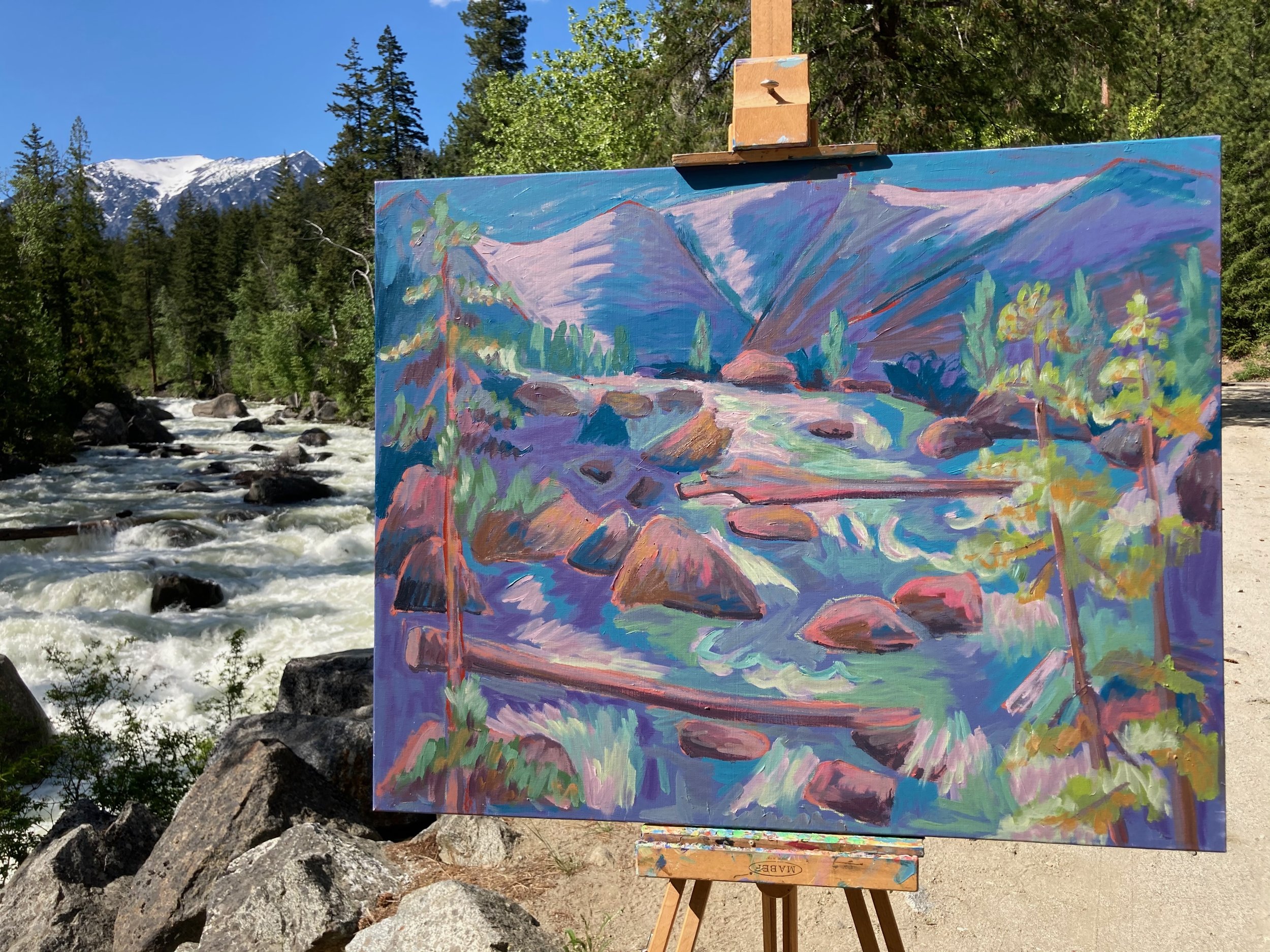 An outdoor painting of a river flowing through a mountainous landscape with trees and rocky terrain, set against a backdrop of snow-capped mountains and a clear blue sky.