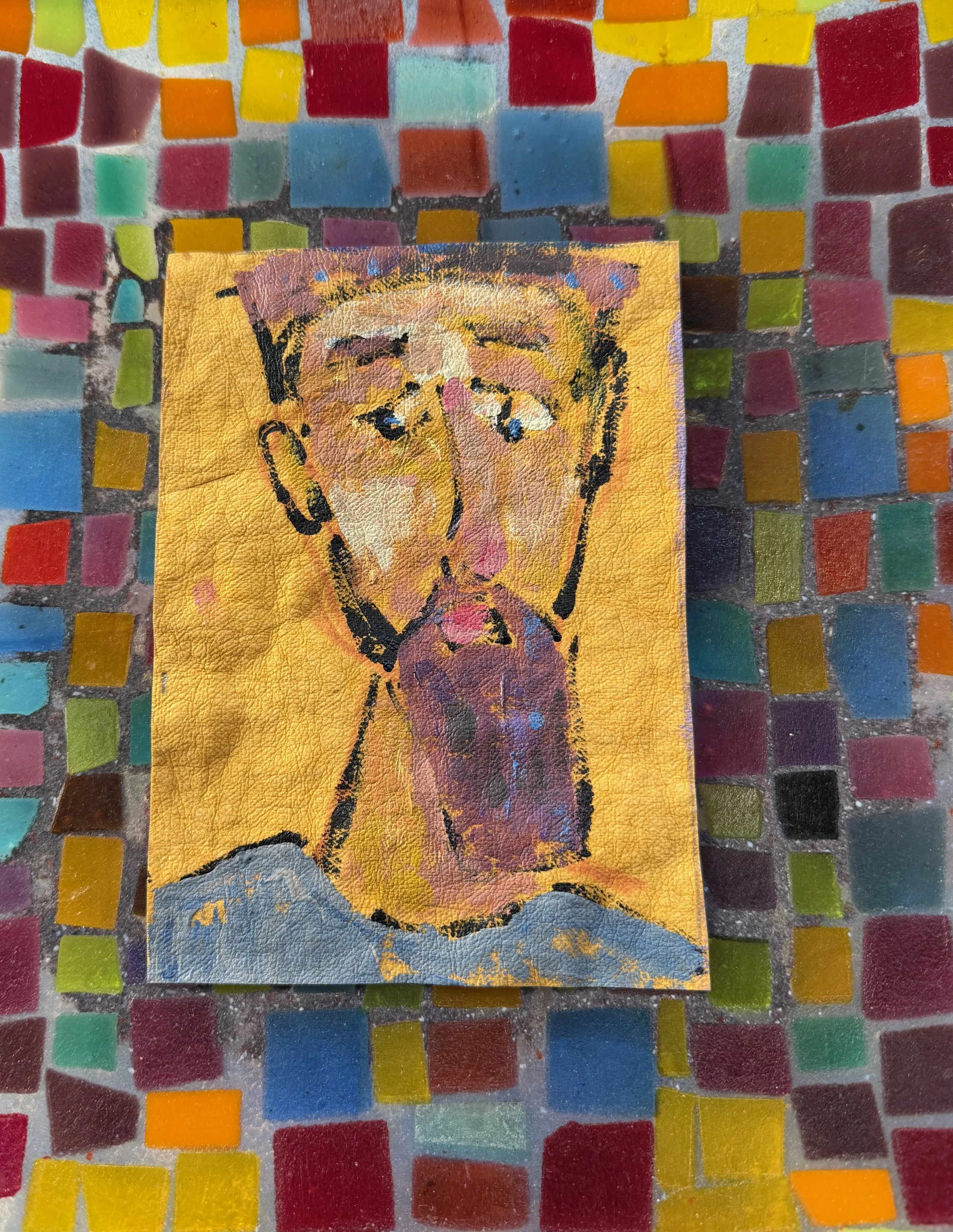 A small painting of a man's face with a beard on a textured yellow background, resting on a multicolored mosaic tiled surface.