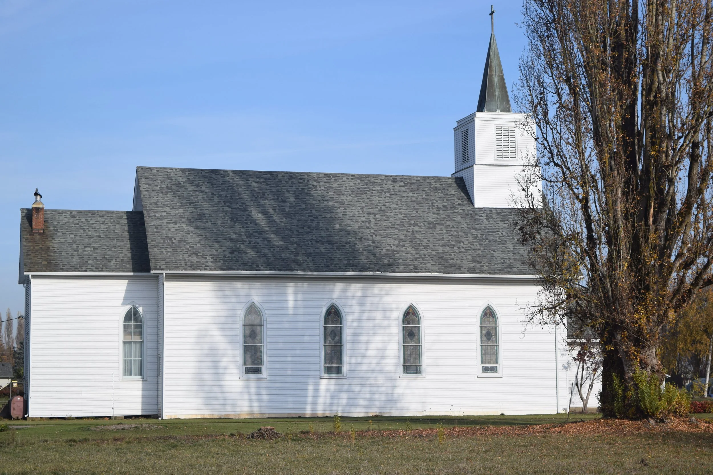 A white church with a tall steeple, arched windows, and a gray shingled roof, situated next to a large leafless tree under a clear blue sky.