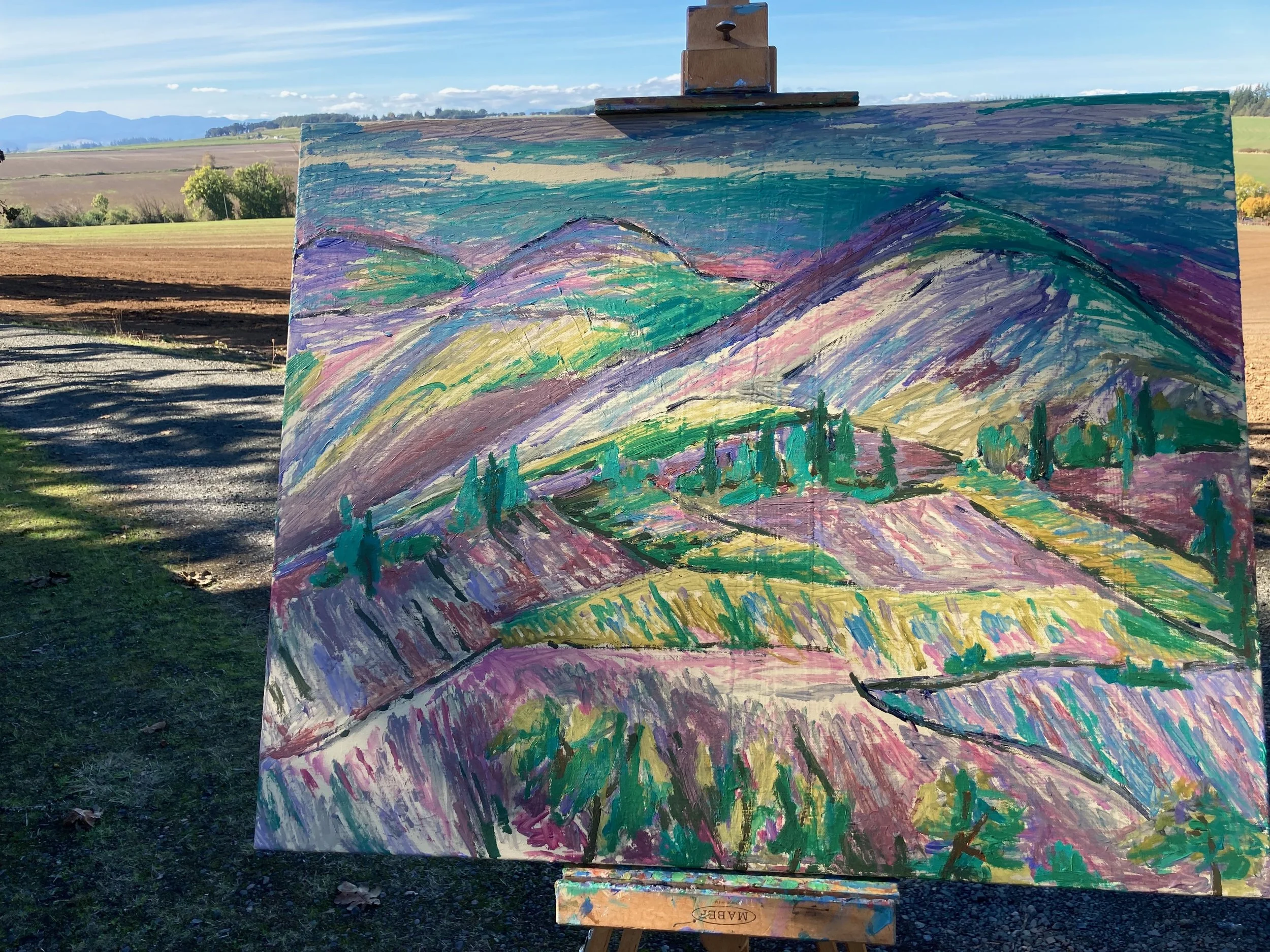 A colorful landscape painting of mountains, hills, and trees, set outdoors on an easel in a rural area with fields, trees, and mountains in the background.