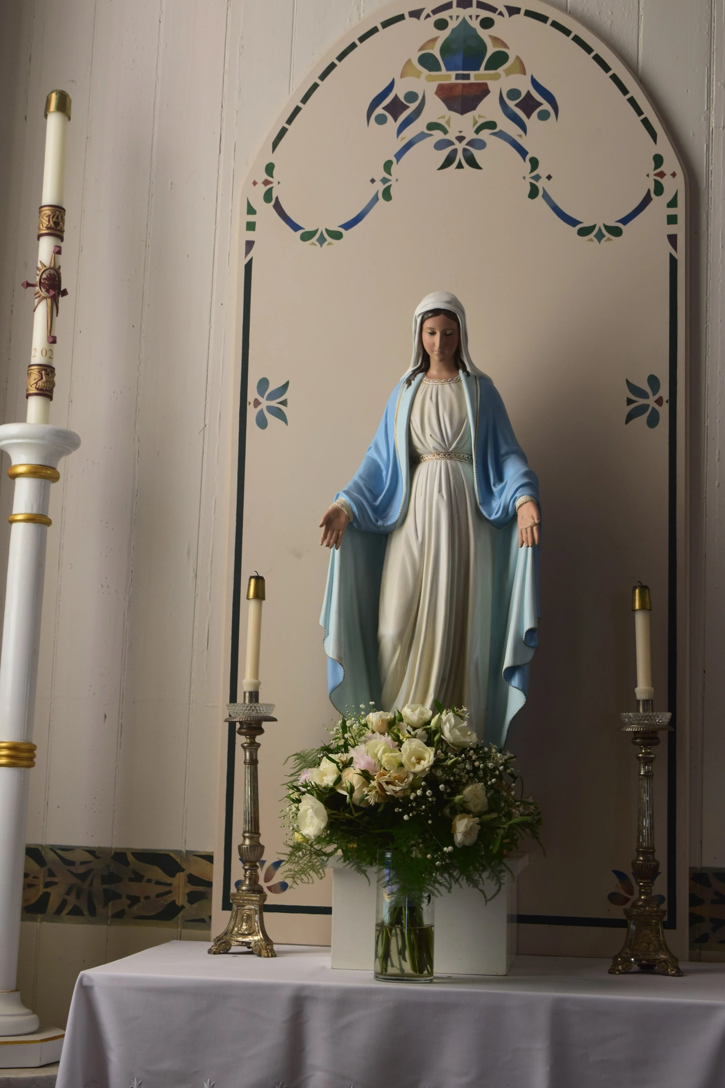 Statue of Virgin Mary dressed in white and blue robes, accented with gold, standing on a white pedestal with a floral arrangement of white and pink roses and greenery in front. The background includes a decorative painted arch with floral and geometr