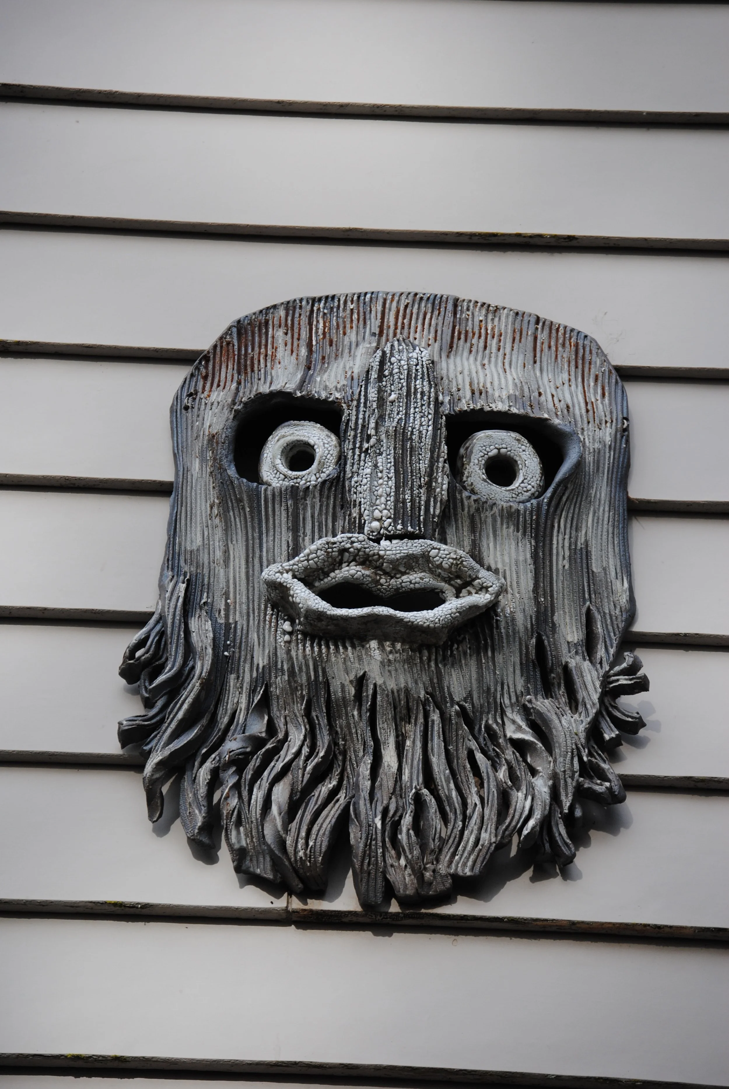 Metal wall art sculpture of a face with a beard, mounted on a white horizontal siding background.
