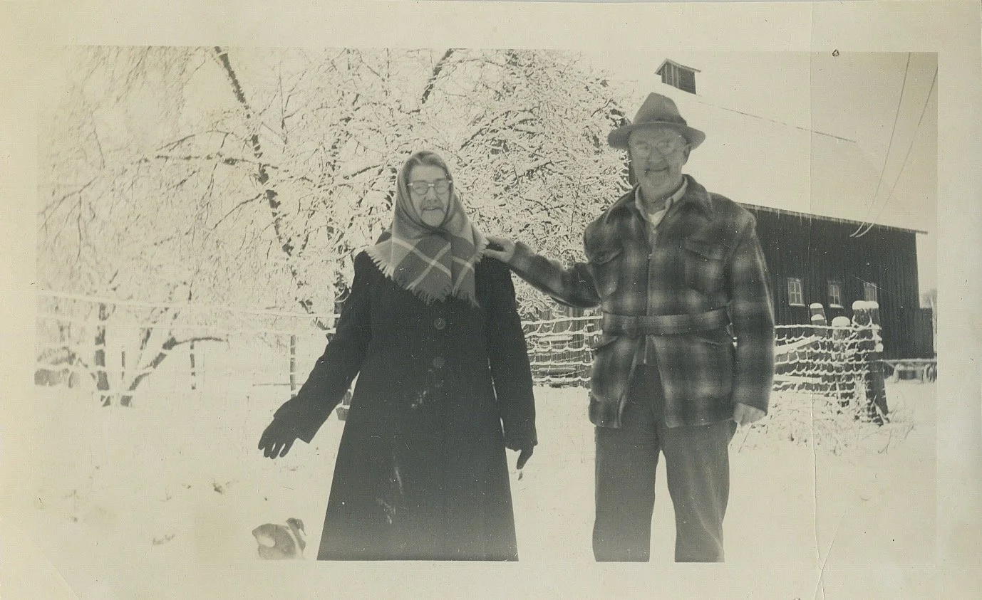 A black-and-white photograph of two people standing outside in a snowy landscape. The woman on the left is wearing a coat, gloves, glasses, and a headscarf, and a small dog is sitting at her feet. The man on the right is wearing a plaid coat, gloves,