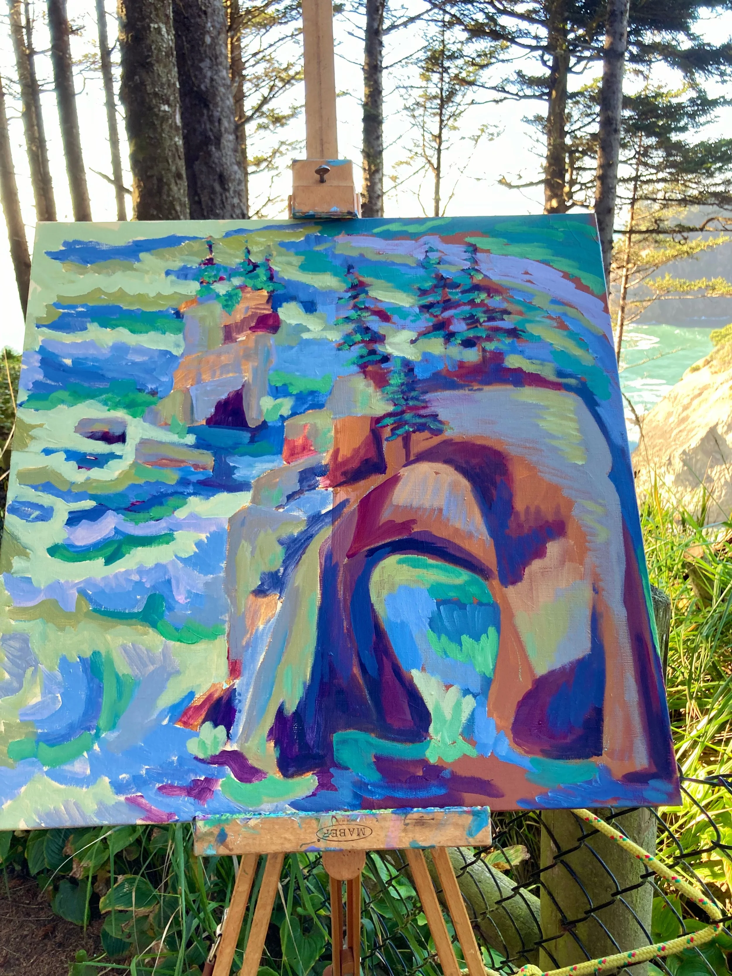 An impressionist painting of a waterfall flowing over rocks surrounded by trees, set on an easel outdoors in a wooded area near a chain-link fence.