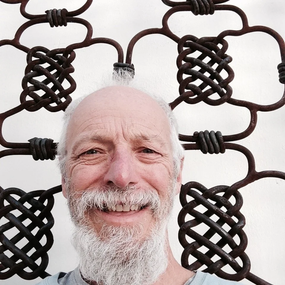 Close-up of an older man with a white beard smiling, standing in front of a decorative metal wall hanging with intricate knot designs.