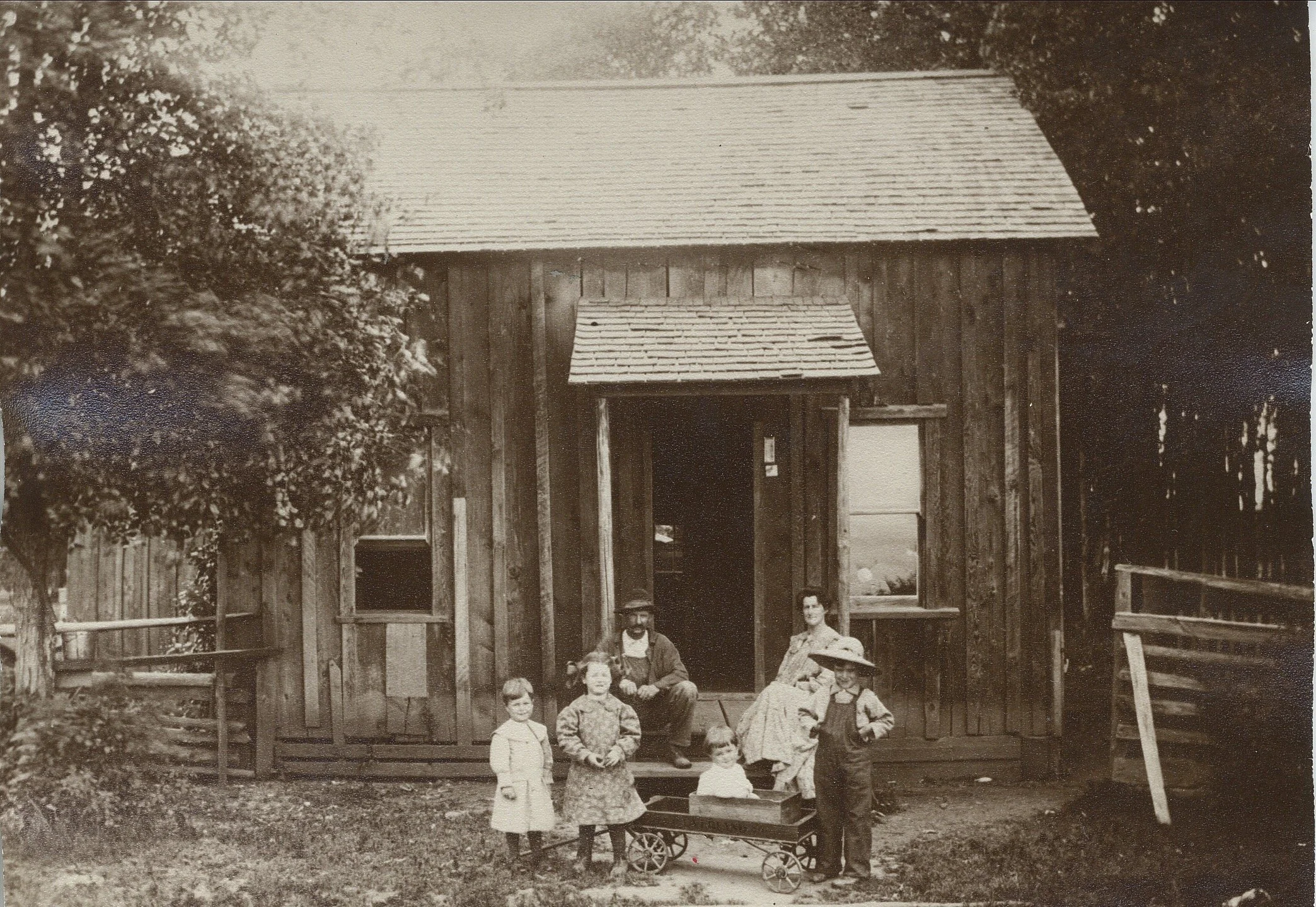 Black and white photo of a family standing outside a wooden house. There are three children, two women, and one man. One of the children is sitting in a small wagon. The house has a simple design with a wooden exterior, a small window, and a porch.