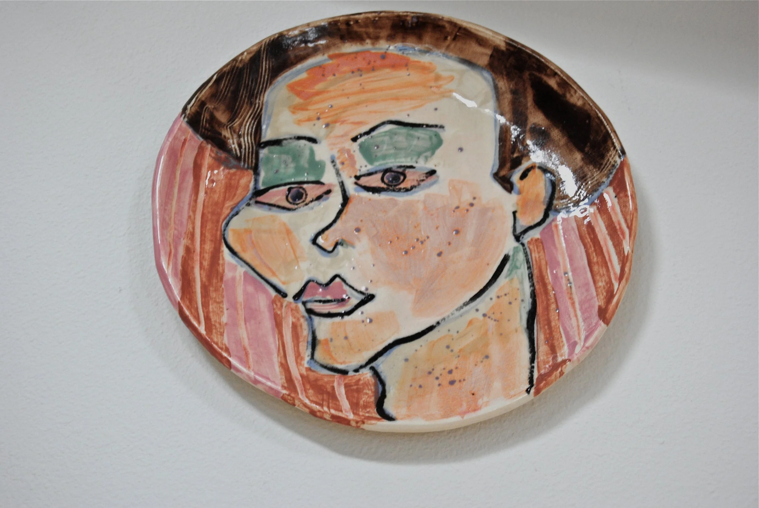 Ceramic plate with a colorful abstract painting of a person's face, featuring green eyes, orange and pink skin tones, and dark hair, mounted on a white wall.