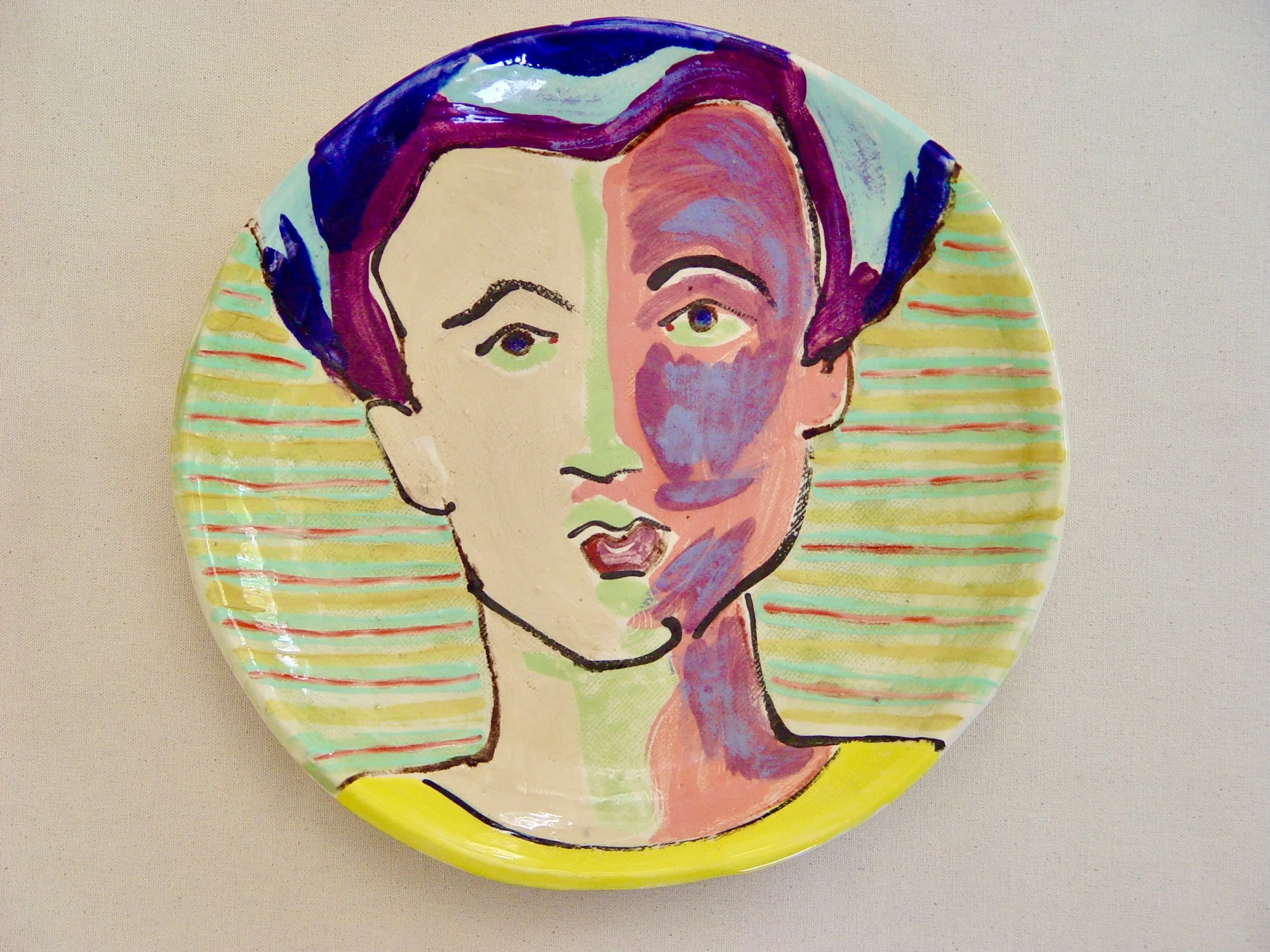 Decorative ceramic plate featuring a stylized portrait of a woman with purple hair and exaggerated facial features, with colorful striped background.