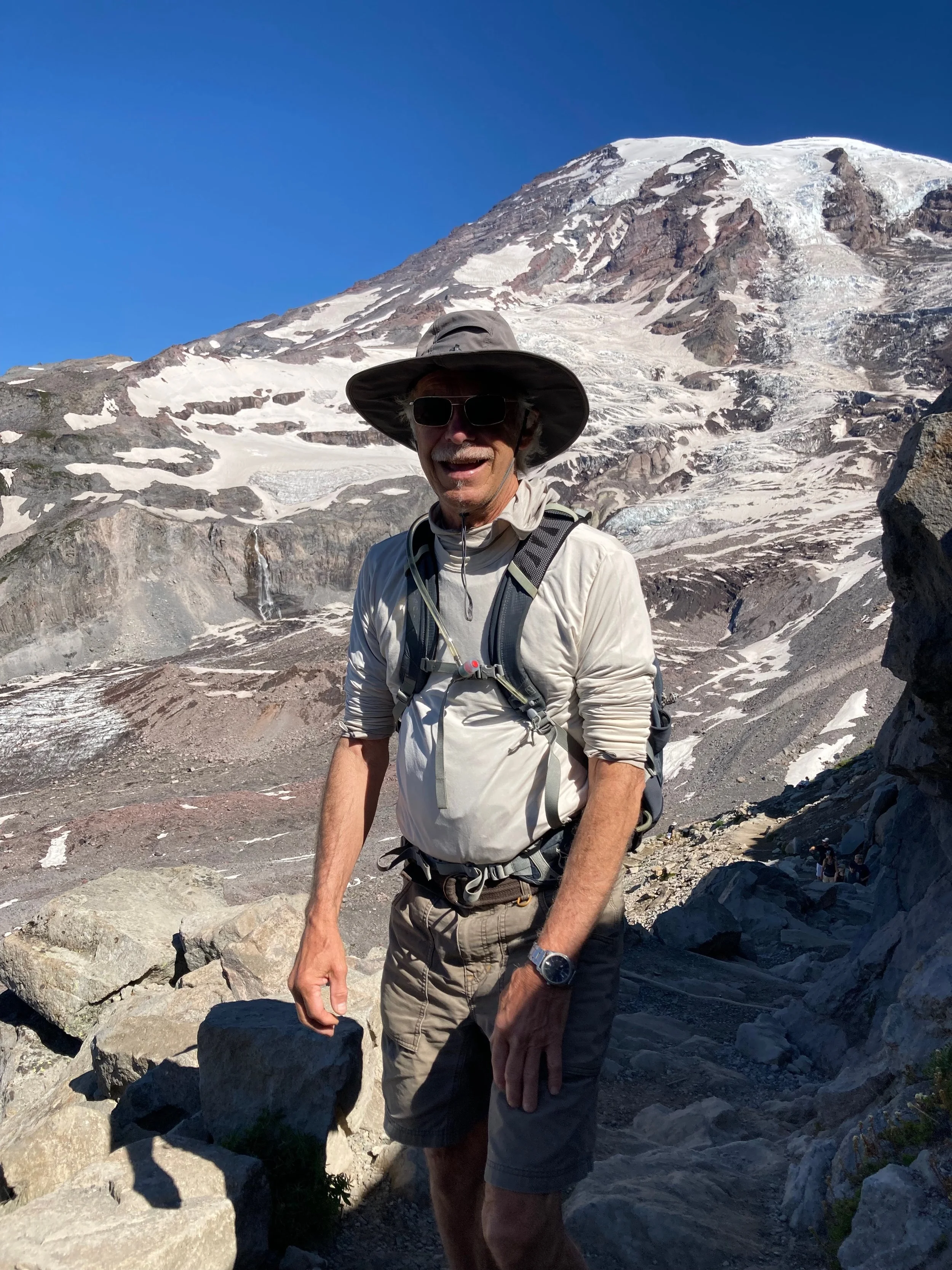 A man wearing a wide-brimmed hat, sunglasses, and outdoor gear, standing on a rocky trail with snow-covered mountain and glacier in the background.