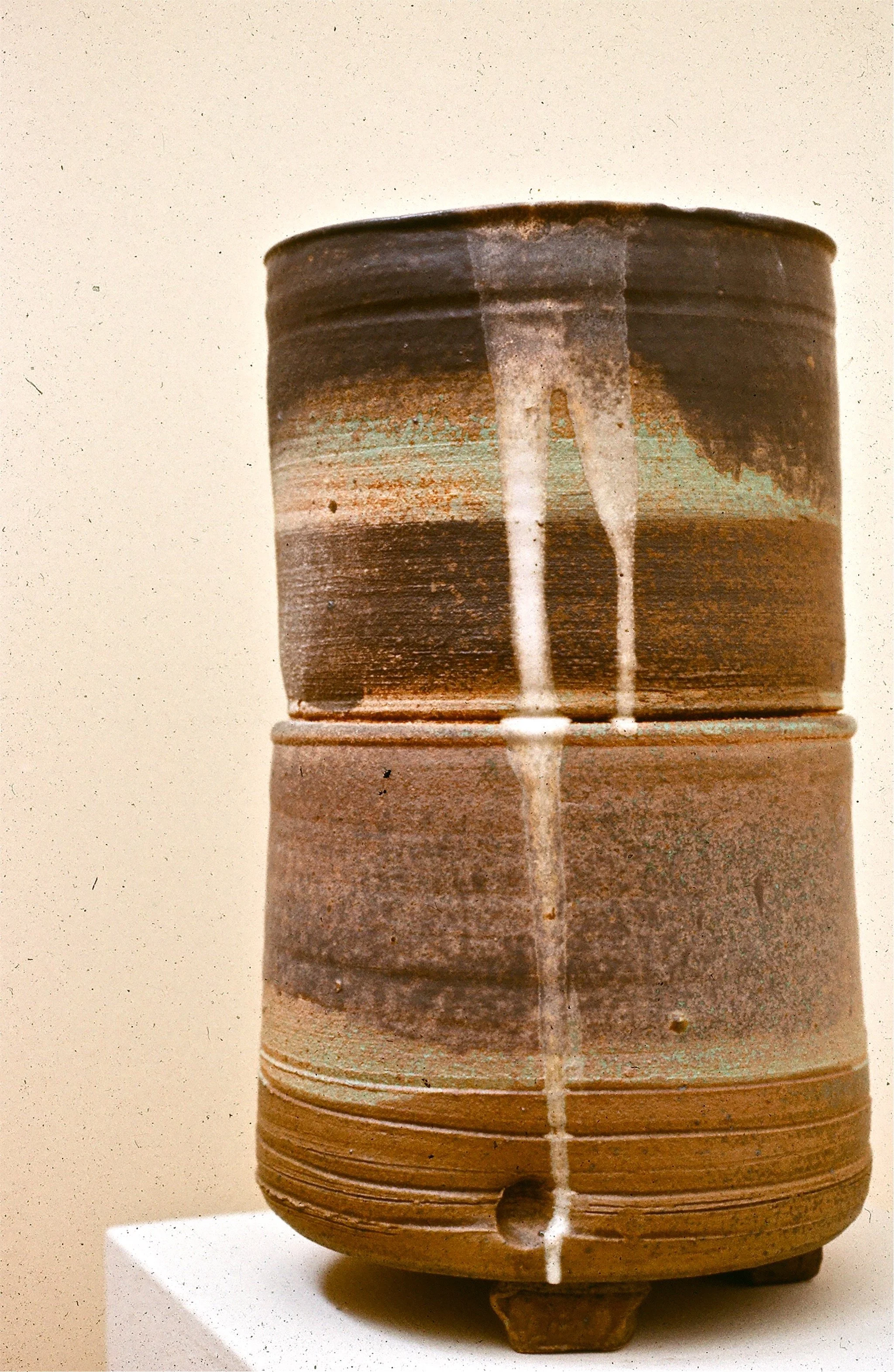 A ceramic vessel with a cylindrical shape, horizontal ridges, and a small hole at the bottom, displaying a layered, natural earth-tone glaze with a white drip pattern running down the center.