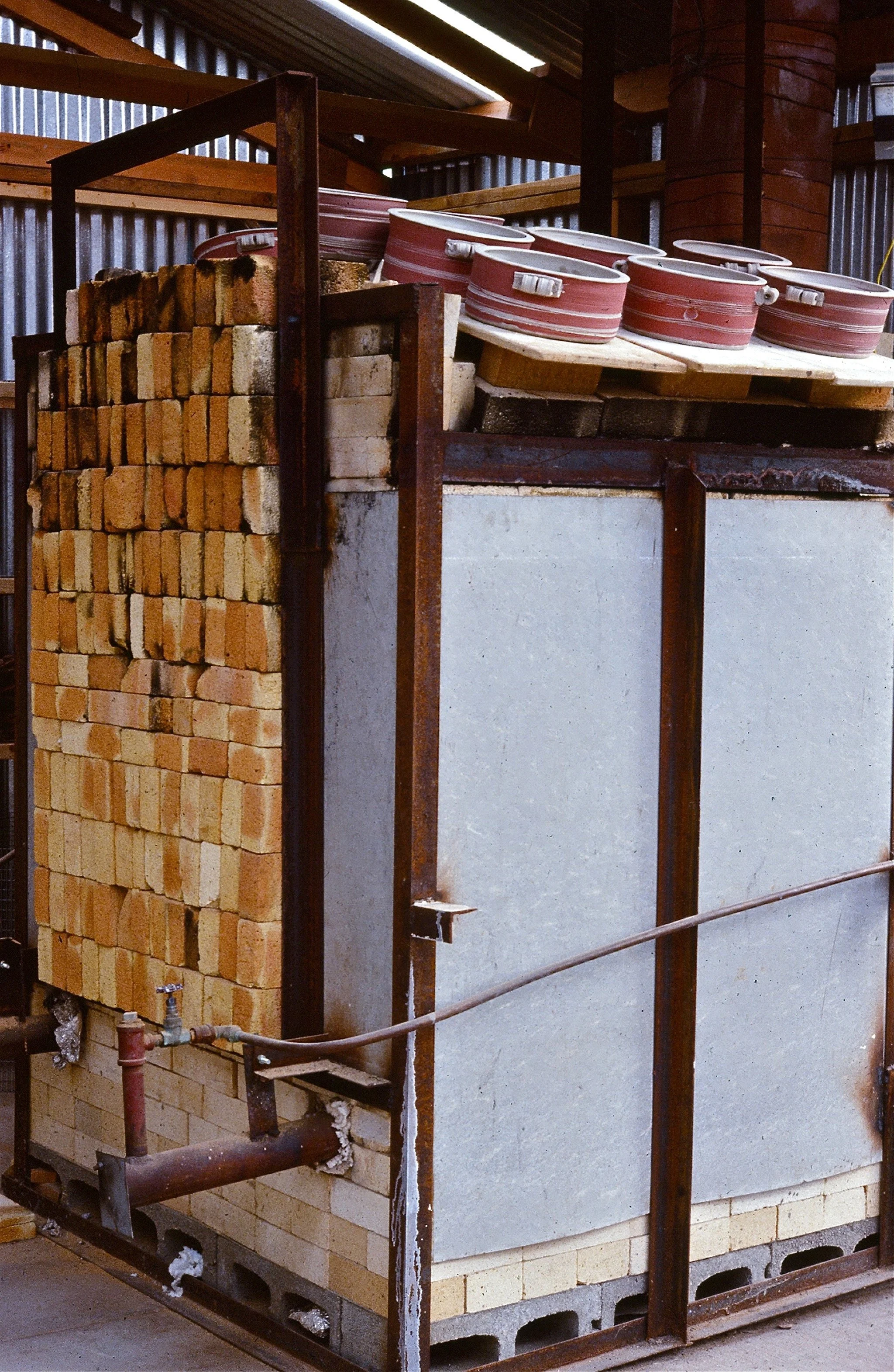 Stacked bricks, red clay pizza oven with white lid, and metal piping inside a workshop or construction site.