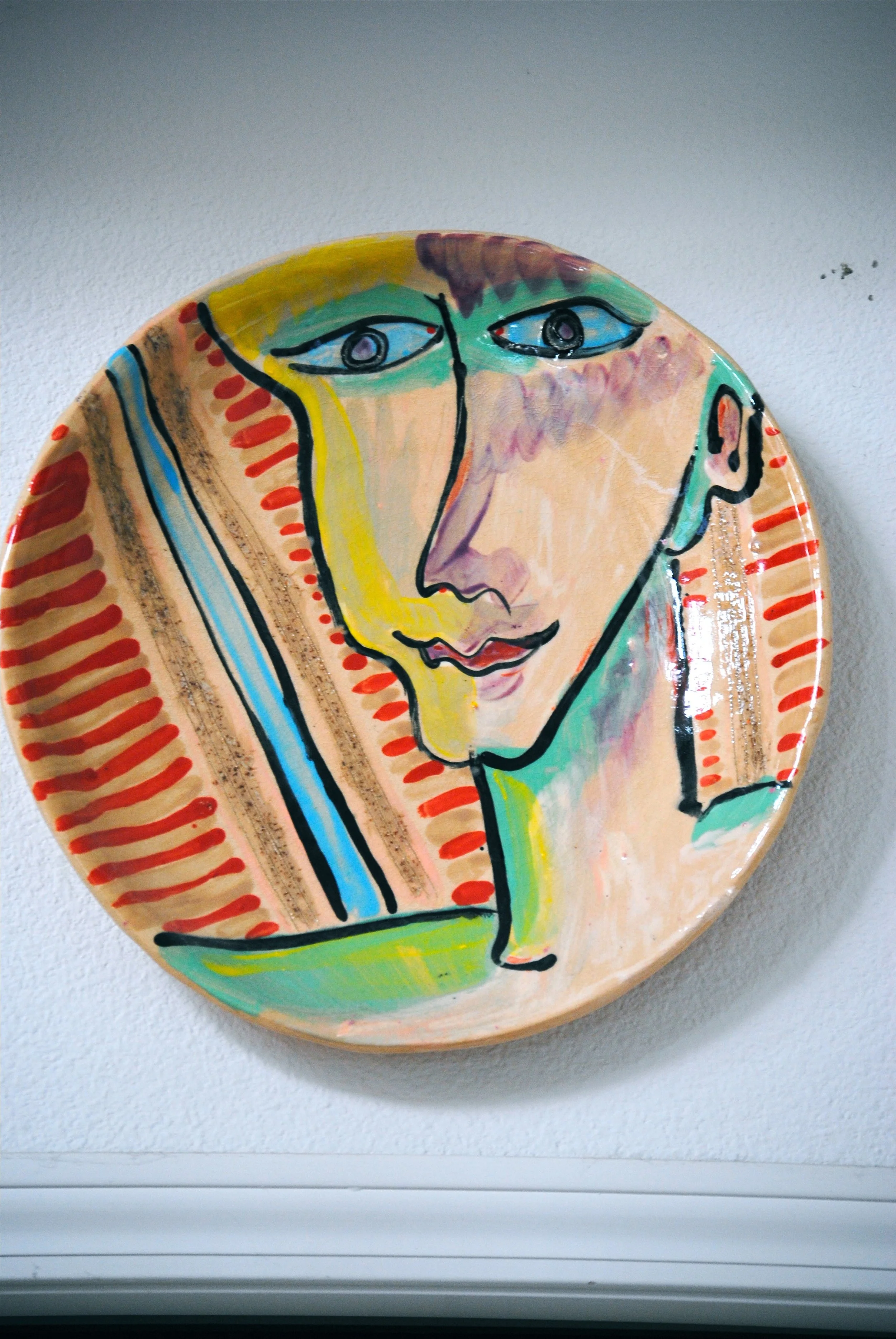 Decorative ceramic plate with an abstract, colorful, face-like painting featuring blue eyes and a gentle smile, mounted on a white wall.