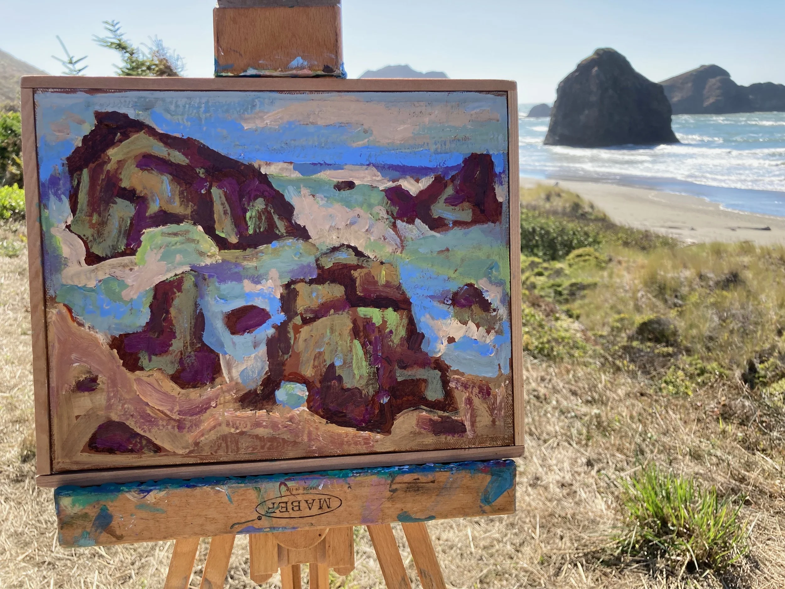 An impressionist oil painting of a rocky coastal landscape with cliffs, water, and sky, displayed on an easel, with a beach, ocean waves, large sea stacks, and clear sky in the background.