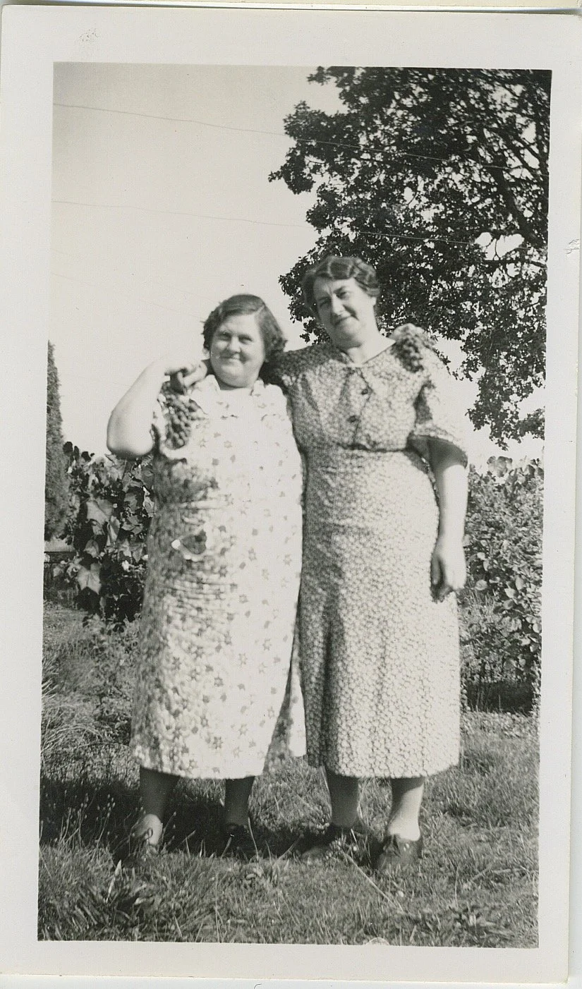Two women standing outdoors, arm-in-arm, smiling, with trees and a clear sky in the background.