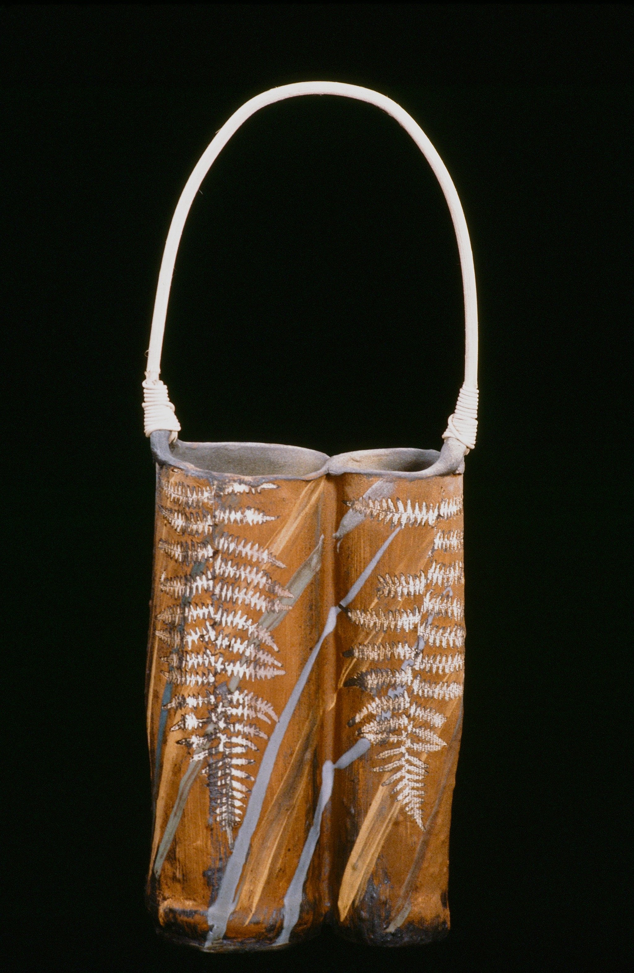 A decorated clay and fiber basket with a white handle, featuring painted fern leaves in white and gray.