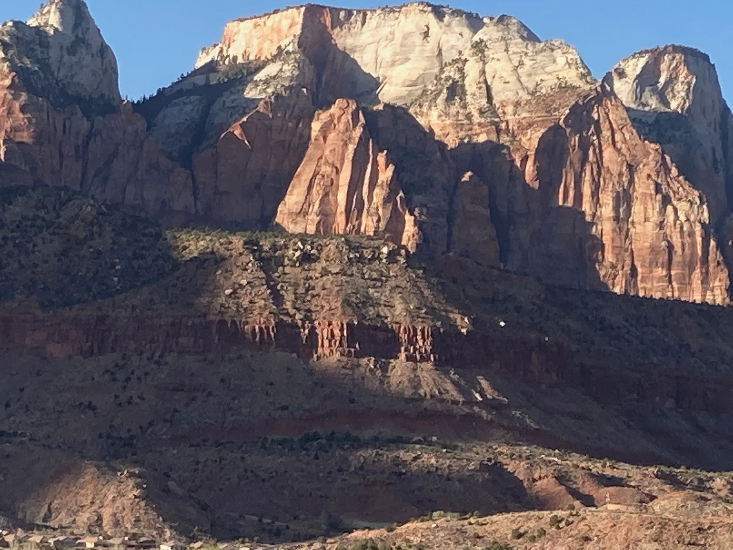 A panoramic view of a mountain range with large, rugged cliffs and a small city or town in the foreground, showing a clear blue sky.
