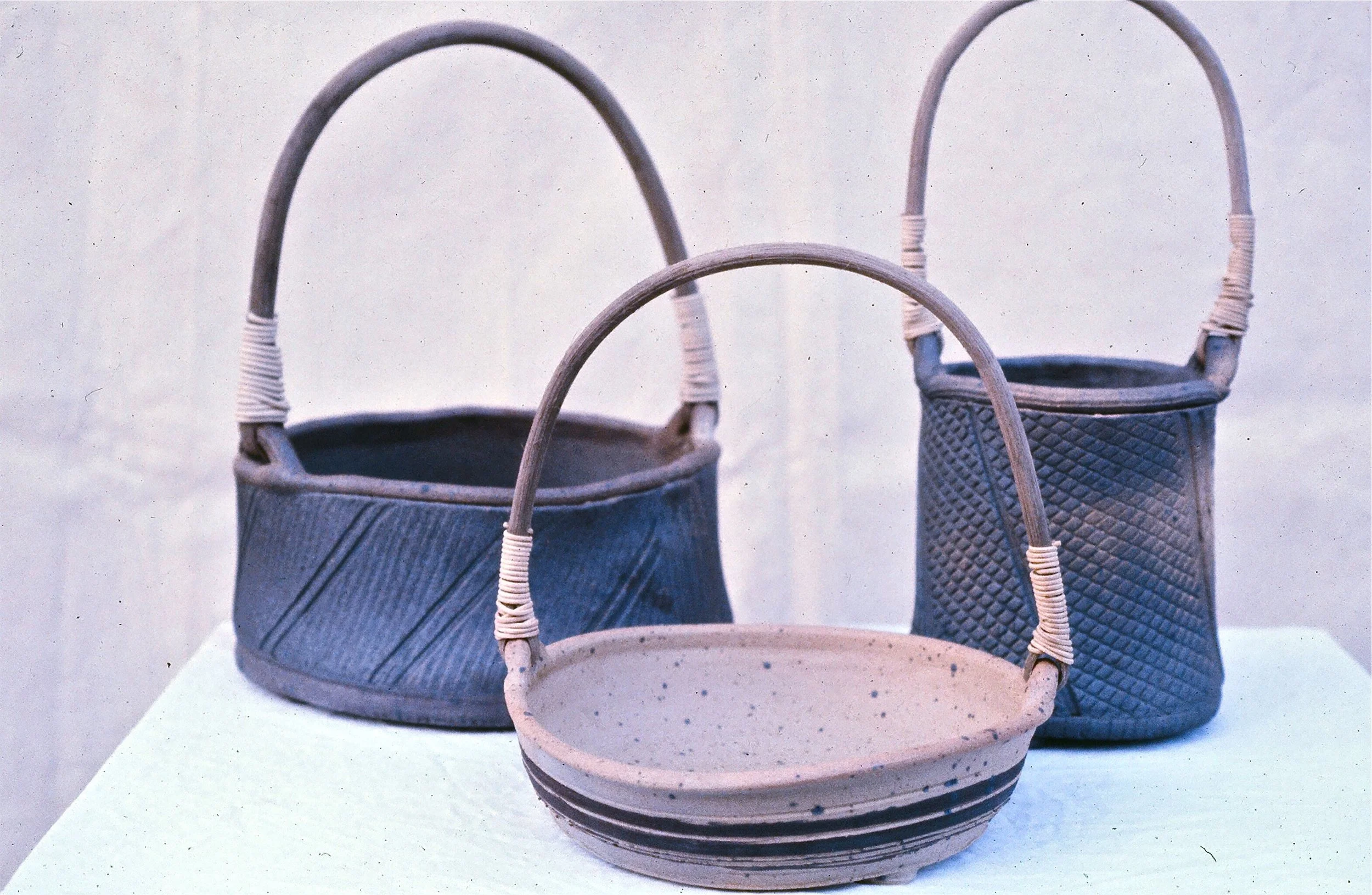Three ceramic baskets with handles, one in front and two in the background, sitting on a white surface against a plain wall.