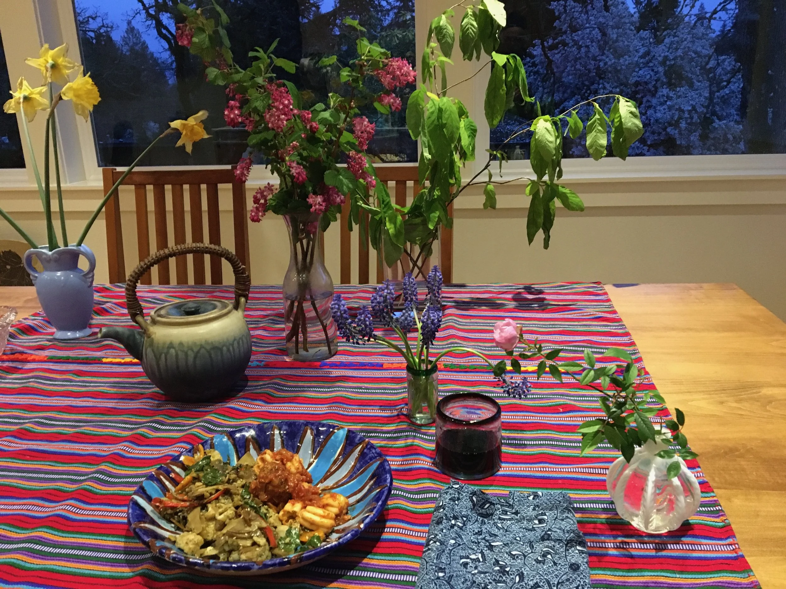 A wooden dining table covered with a colorful striped tablecloth. On the table are several small vases with flowers, a decorative bowl filled with food, a glass of dark beverage, a folded napkin, and a teapot. The background shows large windows with 
