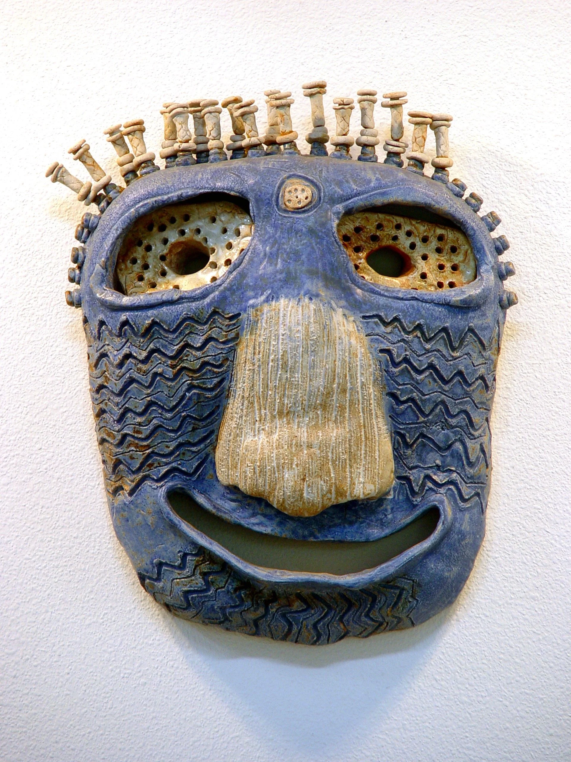 A ceramic mask with a blue face, large eye holes, a textured nose, and a smiling mouth, decorated with small protruding shapes on top and patterned details.