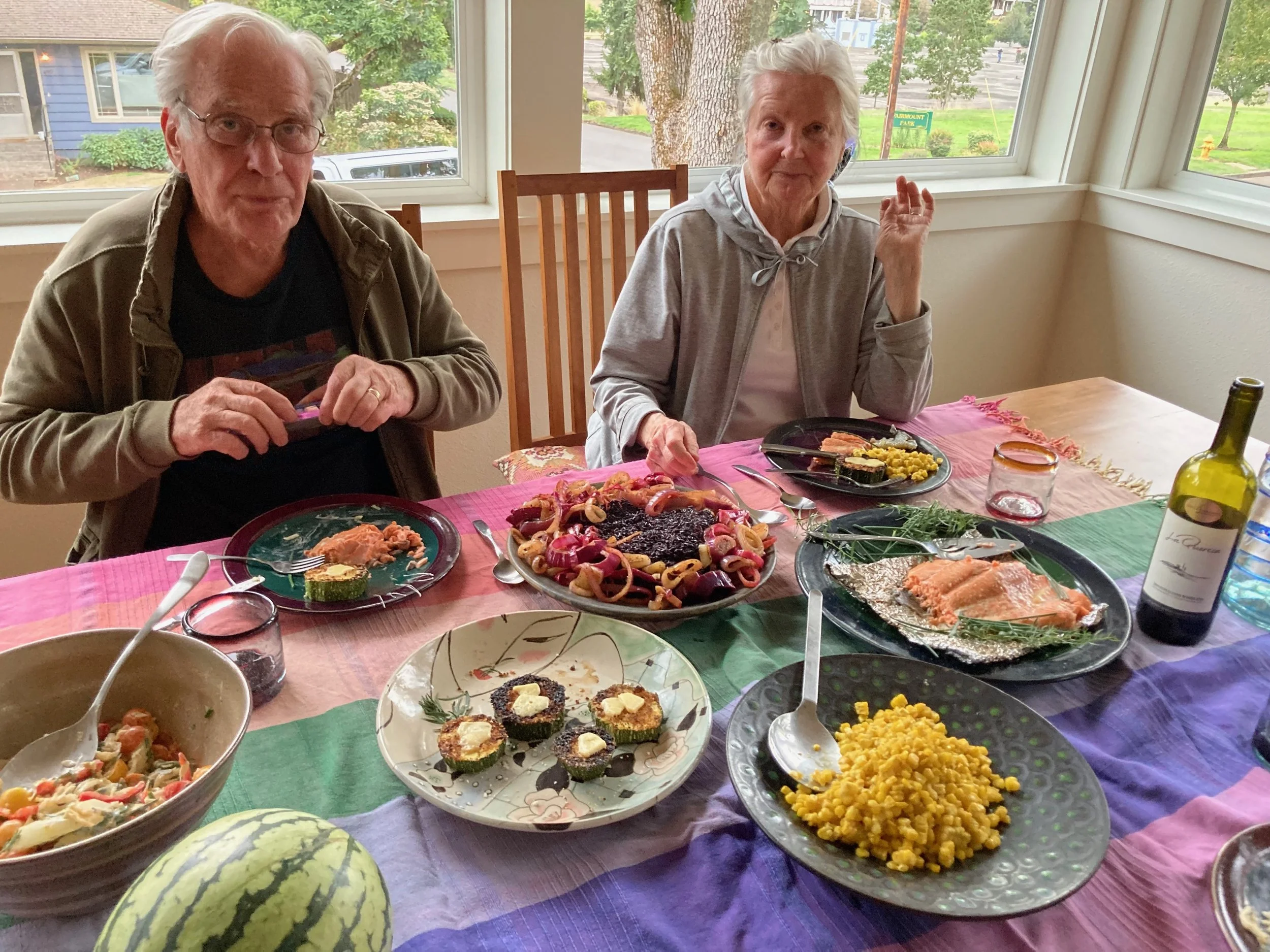 Two elderly women sit at a colorful dining table with various dishes including salmon, salads, corn, and appetizers, in a sunlit room with large windows showing a view of trees and houses outside.