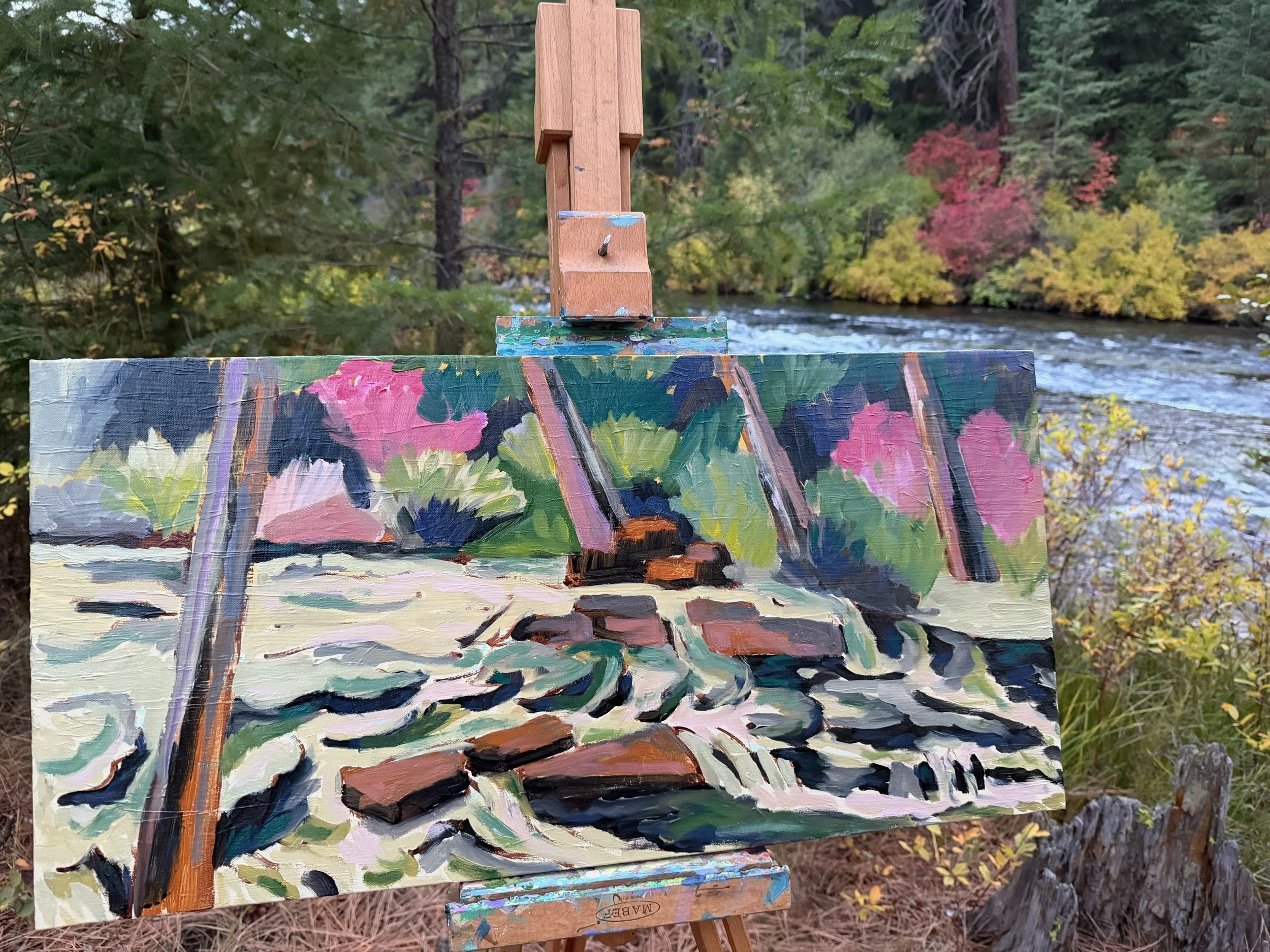 A landscape painting of a flowing stream with rocks, set on an easel outdoors near a river with trees in fall colors in the background.