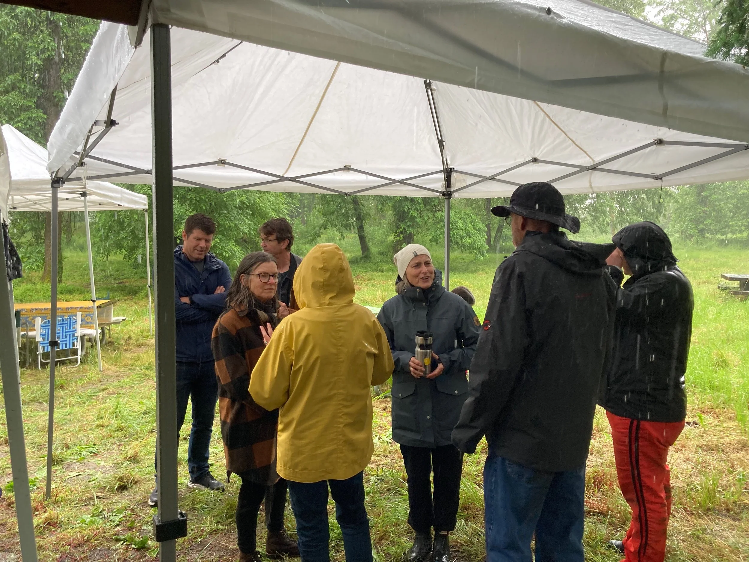 Group of people standing under rain shelter in wet weather, wearing rain jackets, in a grassy outdoor area with trees.