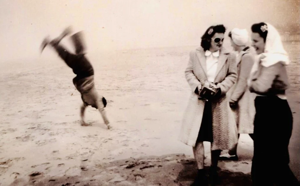 A vintage black-and-white photo of three women standing on the beach, and a person performing a cartwheel in the background.