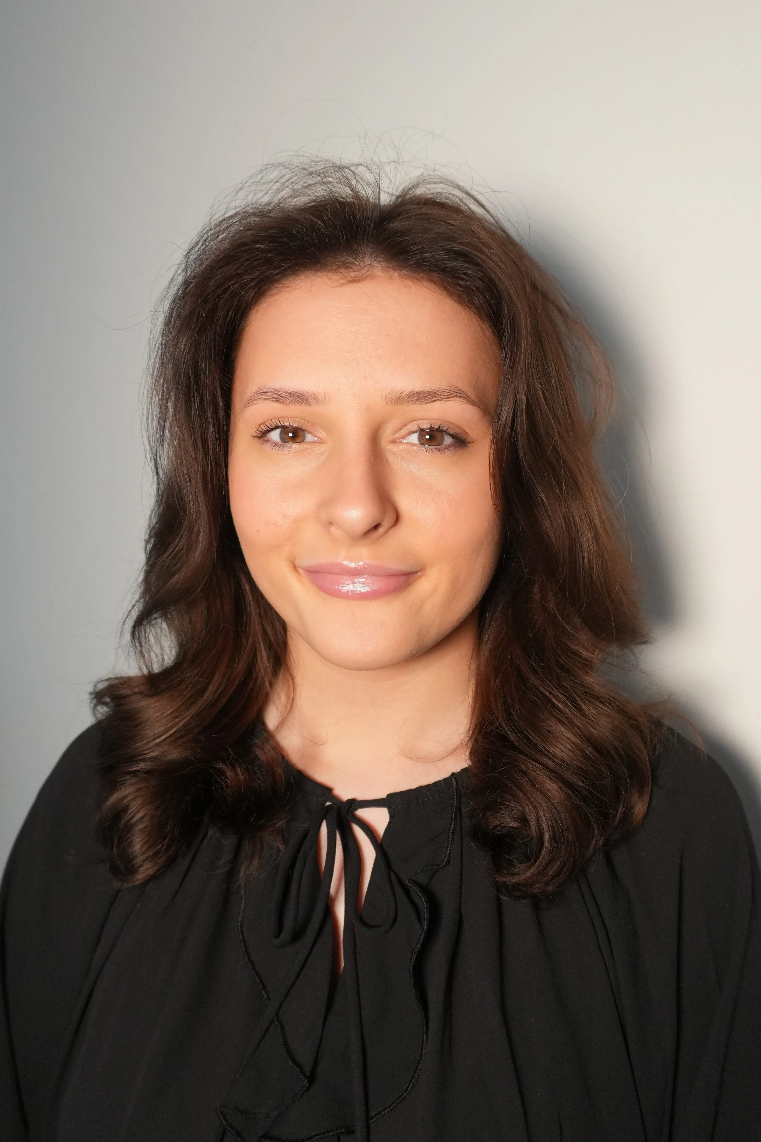 A woman with brown hair smiling and wearing a black top with a bow at the neckline, standing against a neutral background.