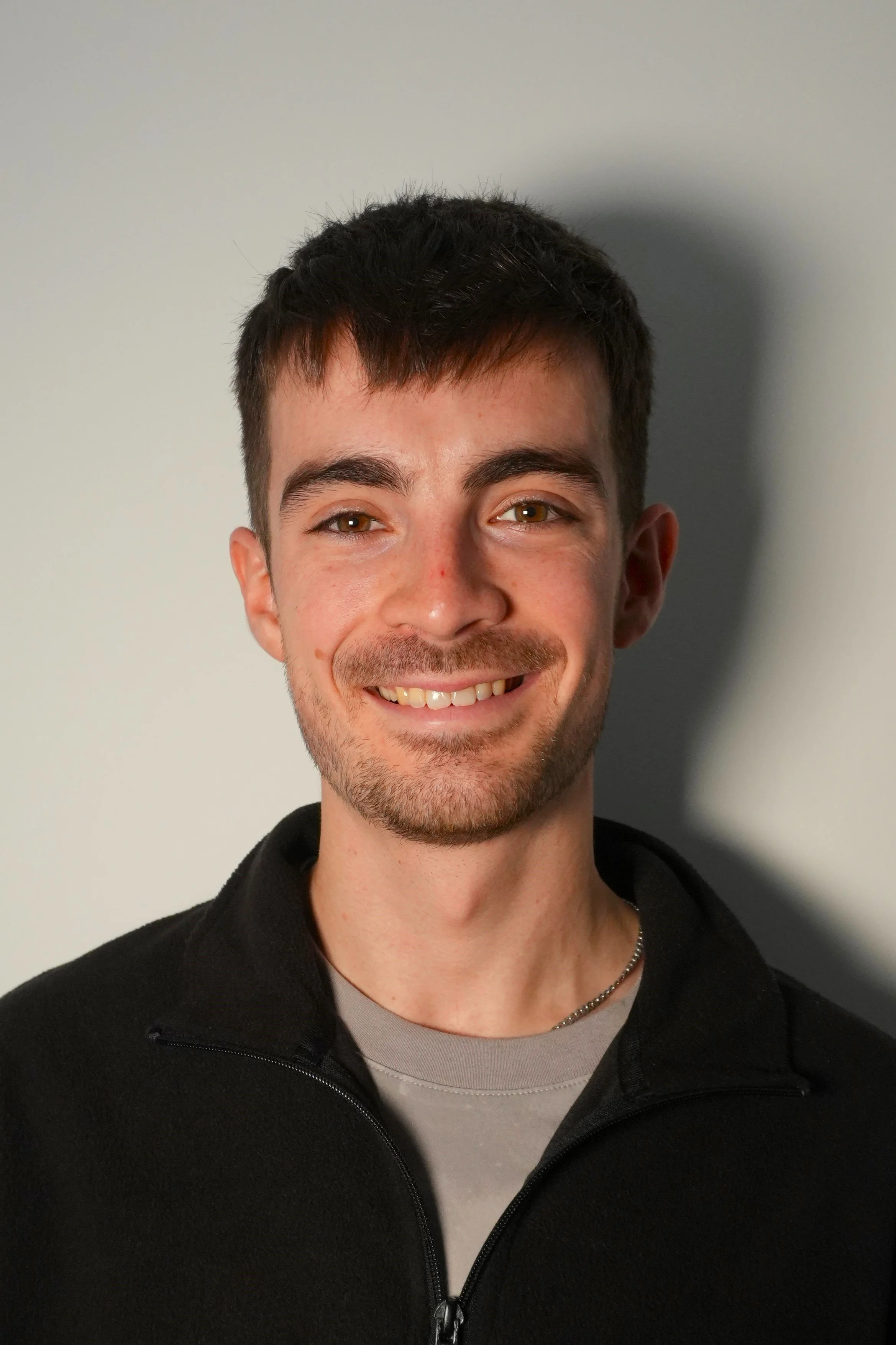 A young man with short dark hair, light beard, and mustache smiling, wearing a black jacket over a gray shirt, standing in front of a plain wall.