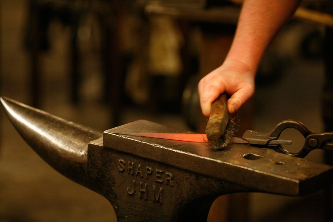 A blacksmith working on a hot metal piece on an anvil with tongs and a brush.