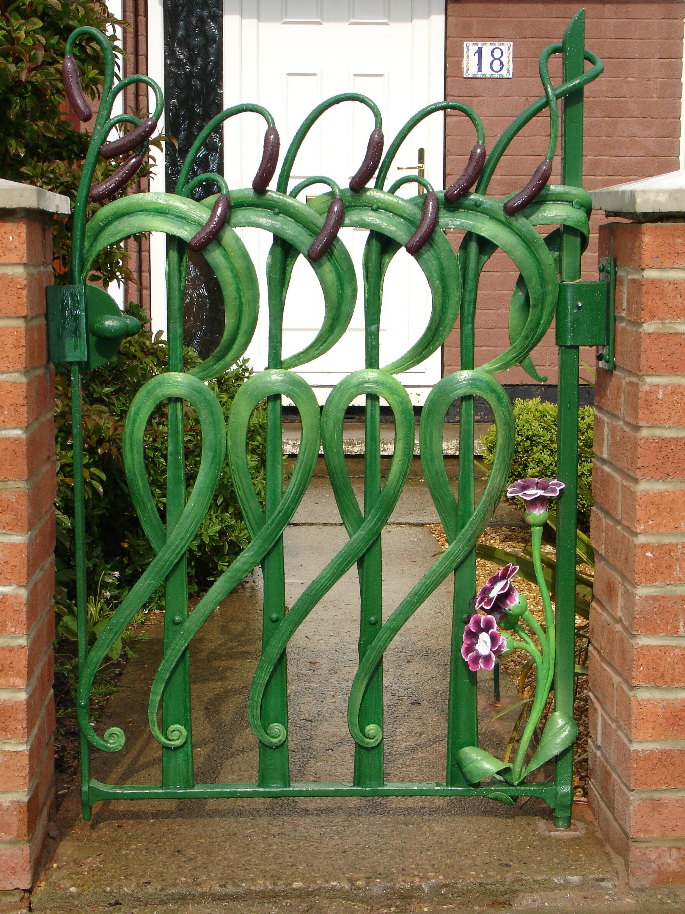 Decorative green garden gate with flower and vine designs, flanked by brick pillars, with house number 18 on a plaque in the background.