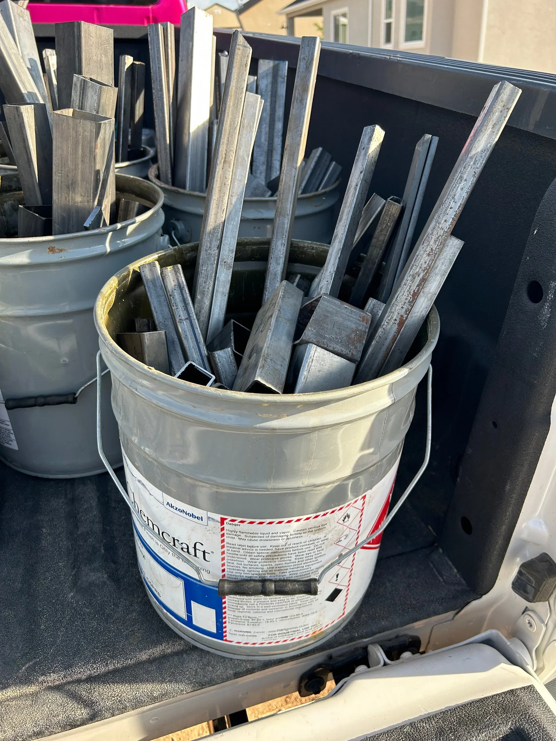 A white bucket filled with metal construction tools and strips, placed on a truck bed with a black plastic lining.