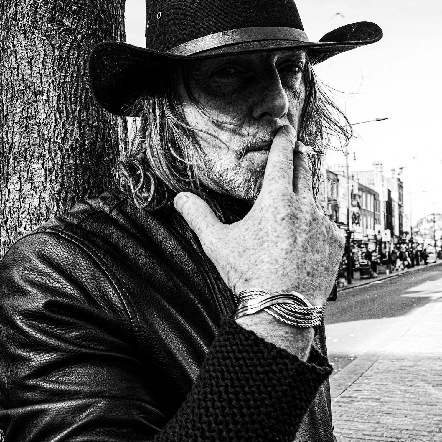 A black and white photo of a man with long, wavy hair, wearing a cowboy hat and leather jacket, smoking a cigarette on a city street.