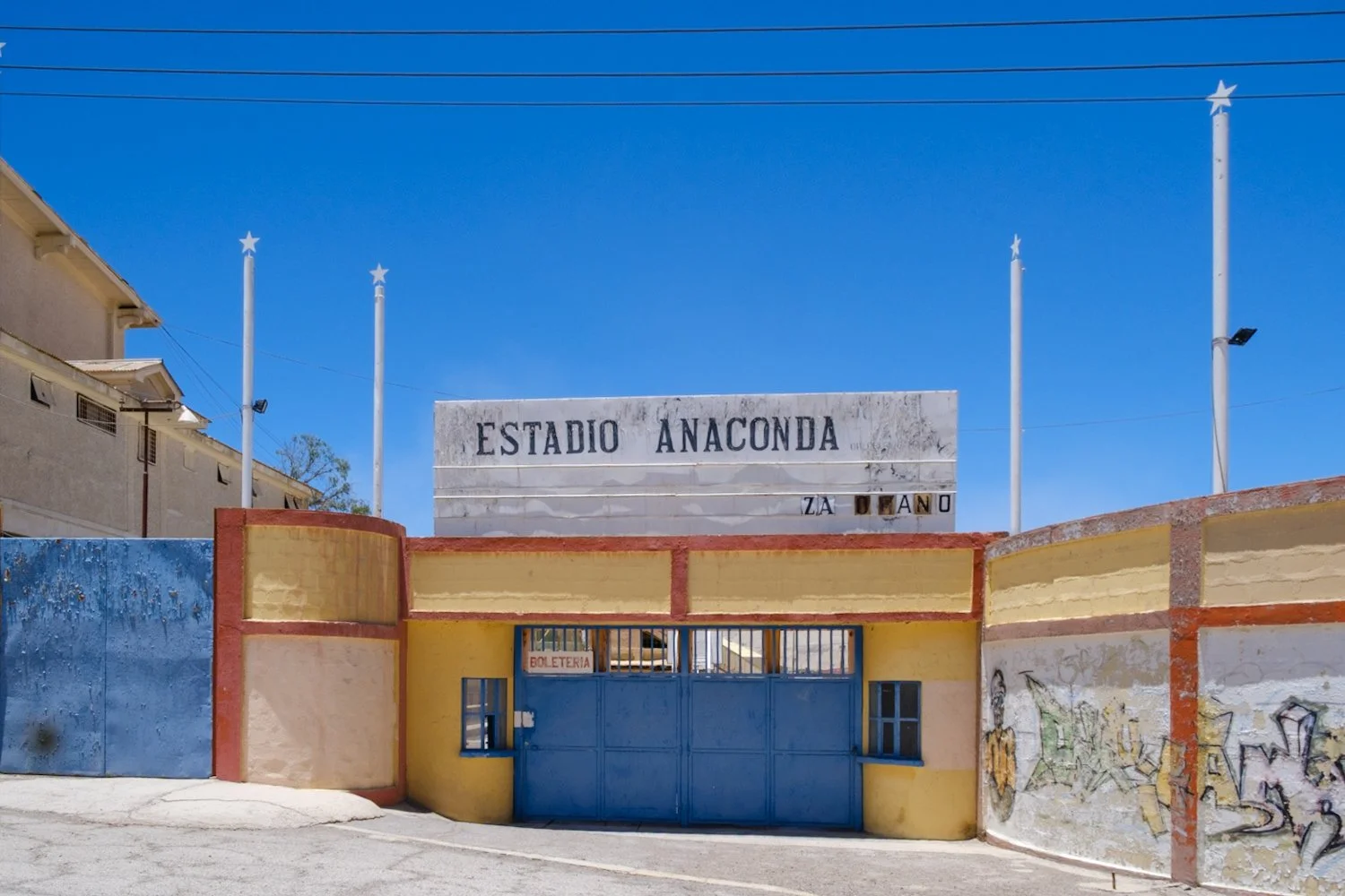 Entrance to Estadio Anaconda with blue gate, yellow and blue walls, graffiti, and four flagpoles against a clear blue sky.
