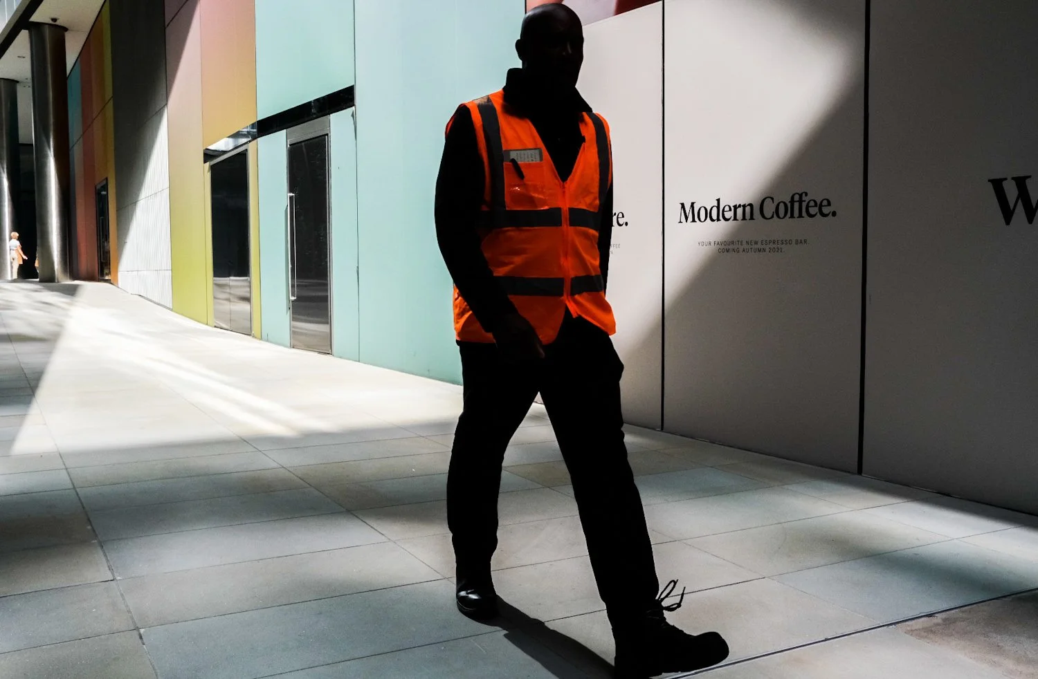 A person wearing an orange safety vest walks through a modern, brightly lit indoor space with colorful wall panels and a large sign that reads "Modern Coffee."