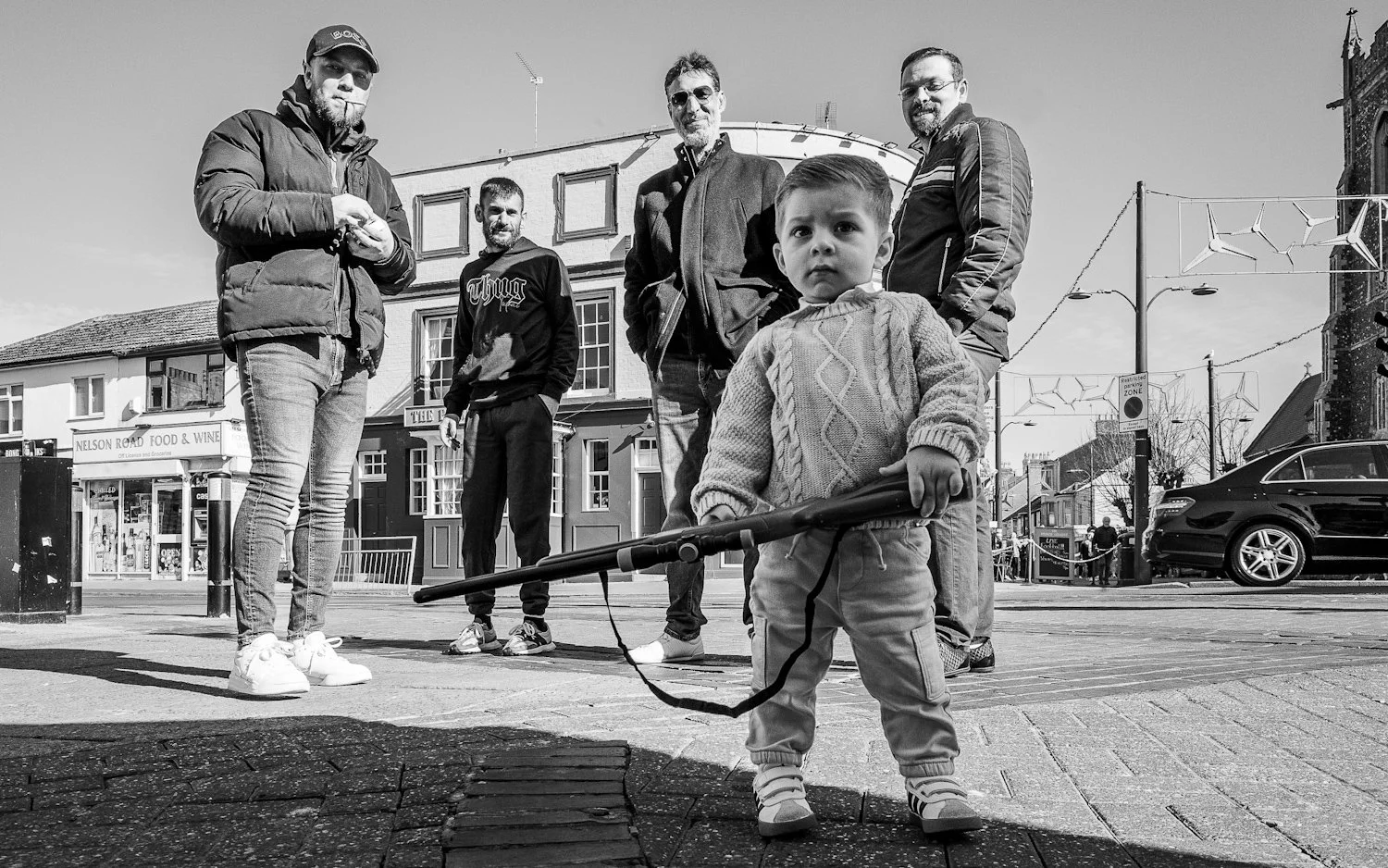 A young boy in a sweater and pants holding a toy rifle, standing in front of a group of five men on a city street in black and white.