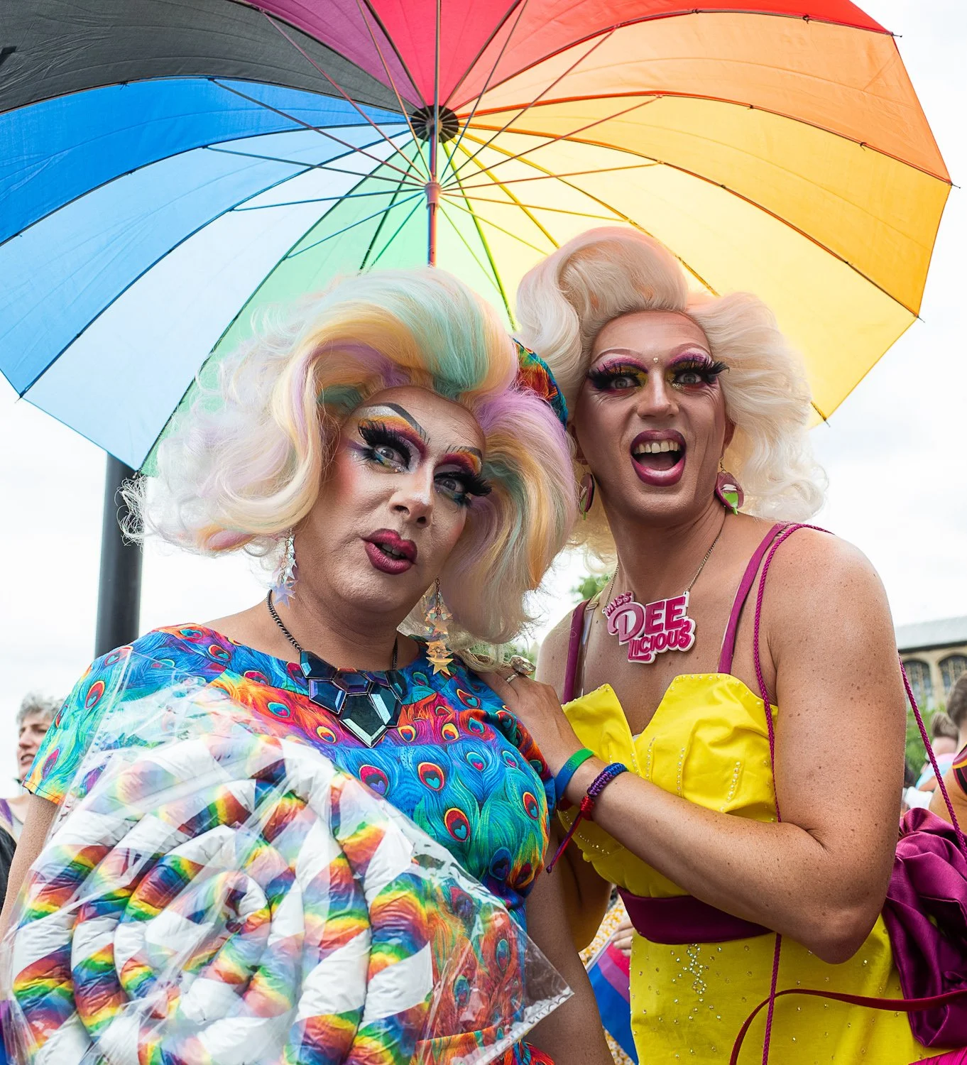 Two individuals dressed in colorful Pride-themed outfits and makeup, standing under a rainbow umbrella at an outdoor Pride event.