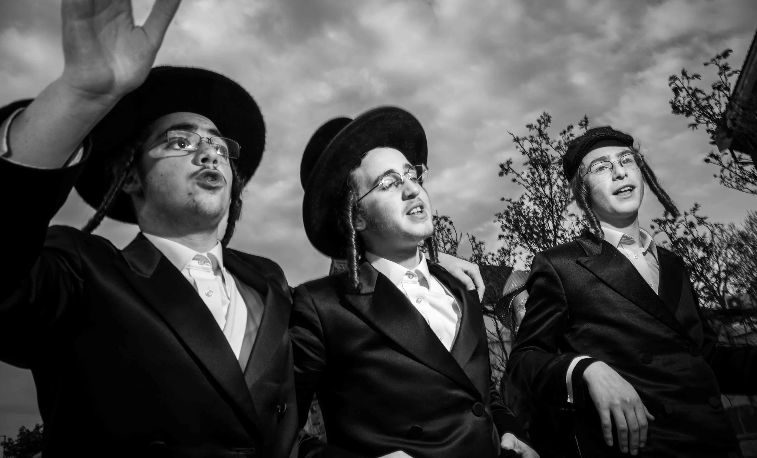 Three young men dressed in academic robes and caps, standing outdoors with arms around each other, looking into the distance and raising a hand, possibly during a graduation or academic ceremony, with a cloudy sky and trees in the background.